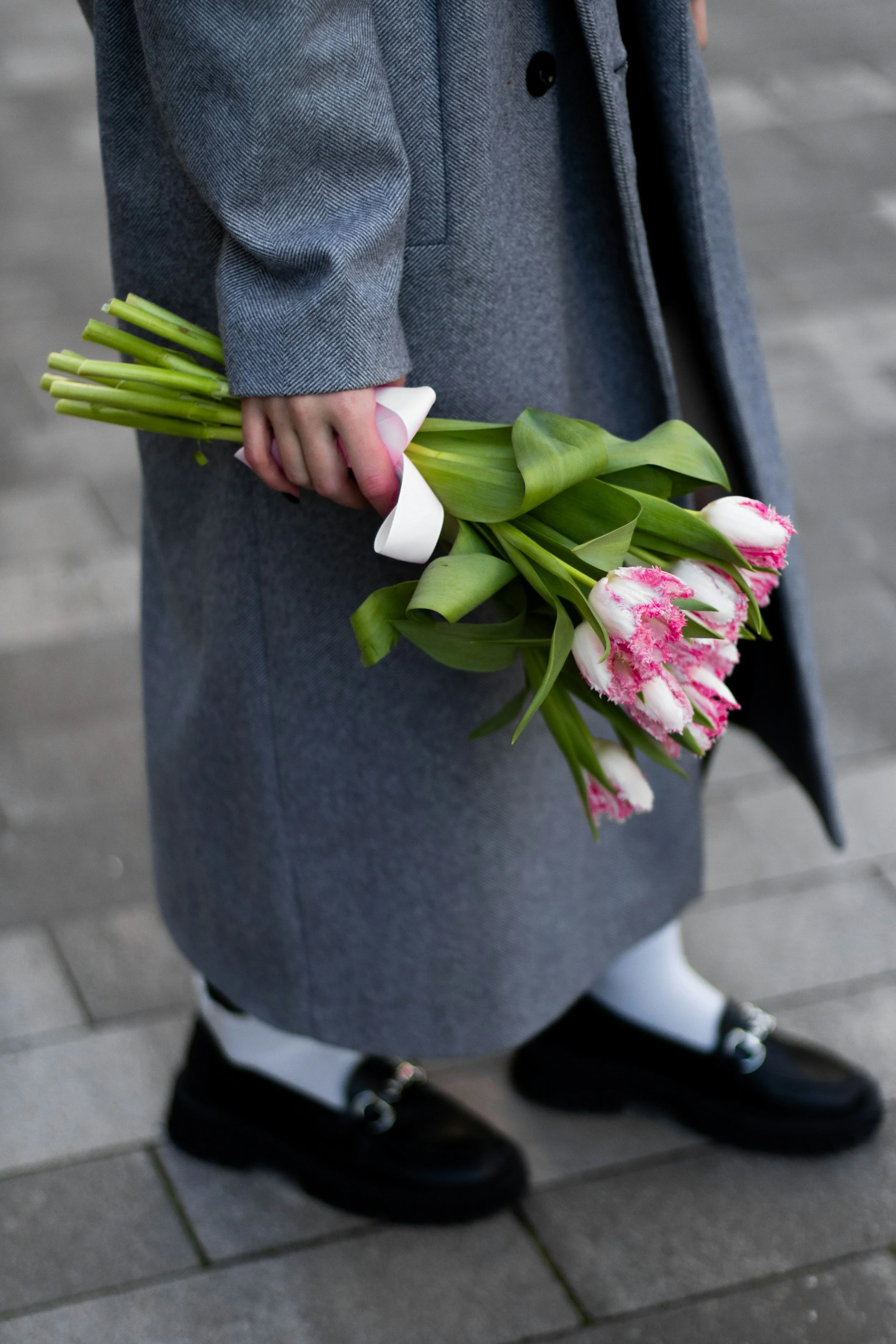 Person wearing a gray coat and black shoes holding a bouquet of pink and white tulips with green leaves.
