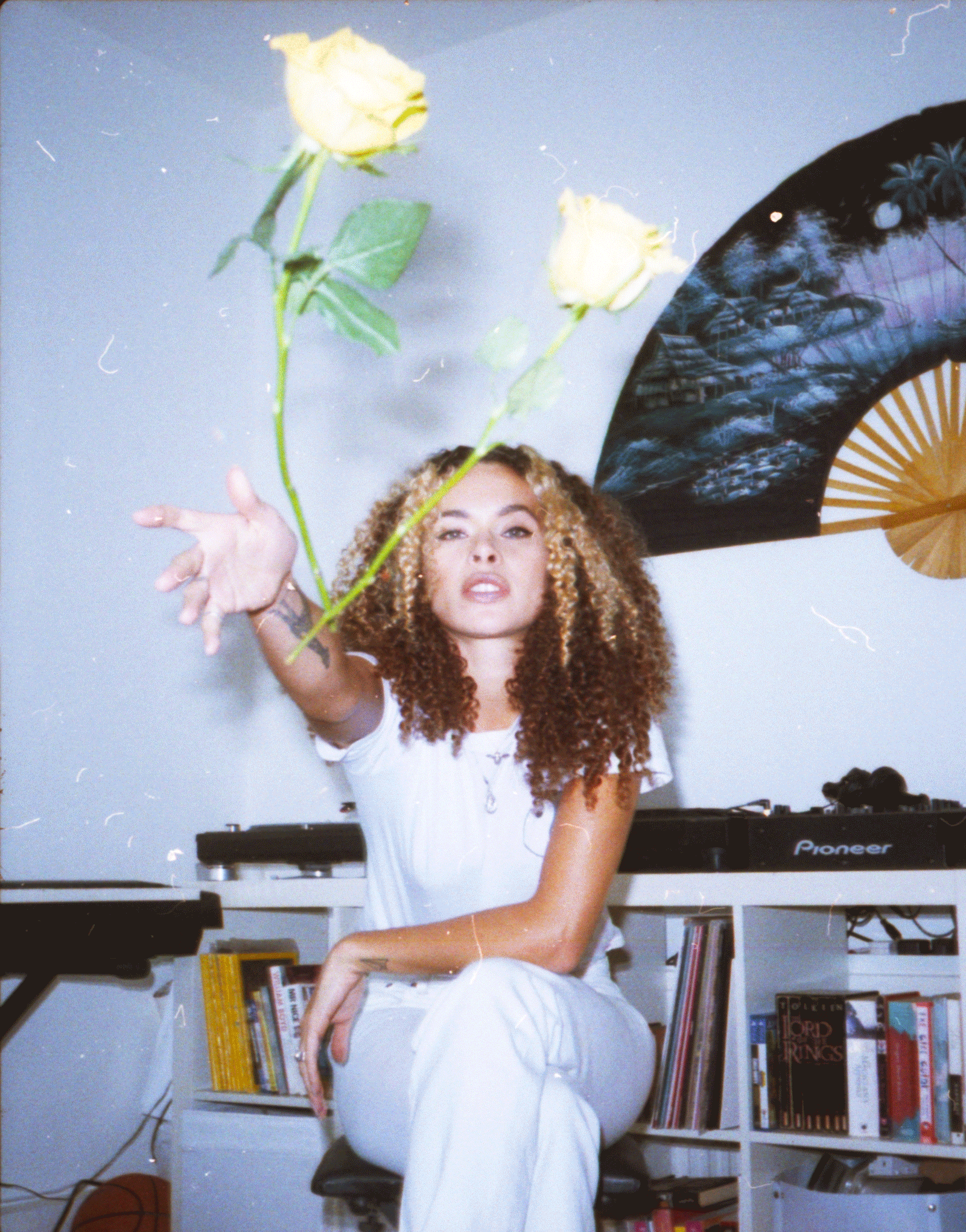 A woman with curly hair sitting on a stool in a room with a white bookshelf filled with books behind her, a DJ turntable, and a decorative umbrella on the wall. She is reaching out towards the camera with three yellow roses in the foreground.