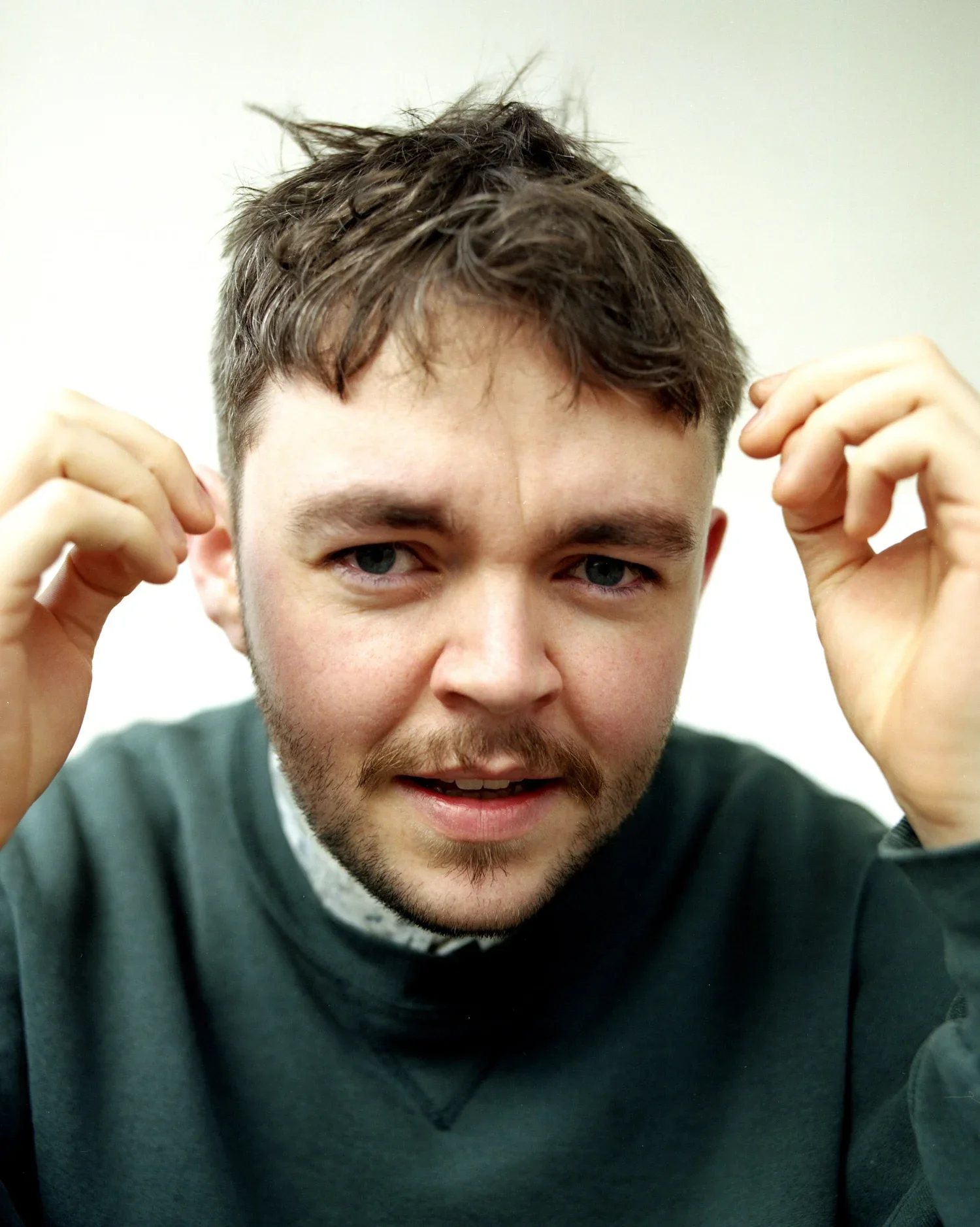 A young man with tousled brown hair and light facial hair is holding his hands near his head and making a slightly playful or confused expression.