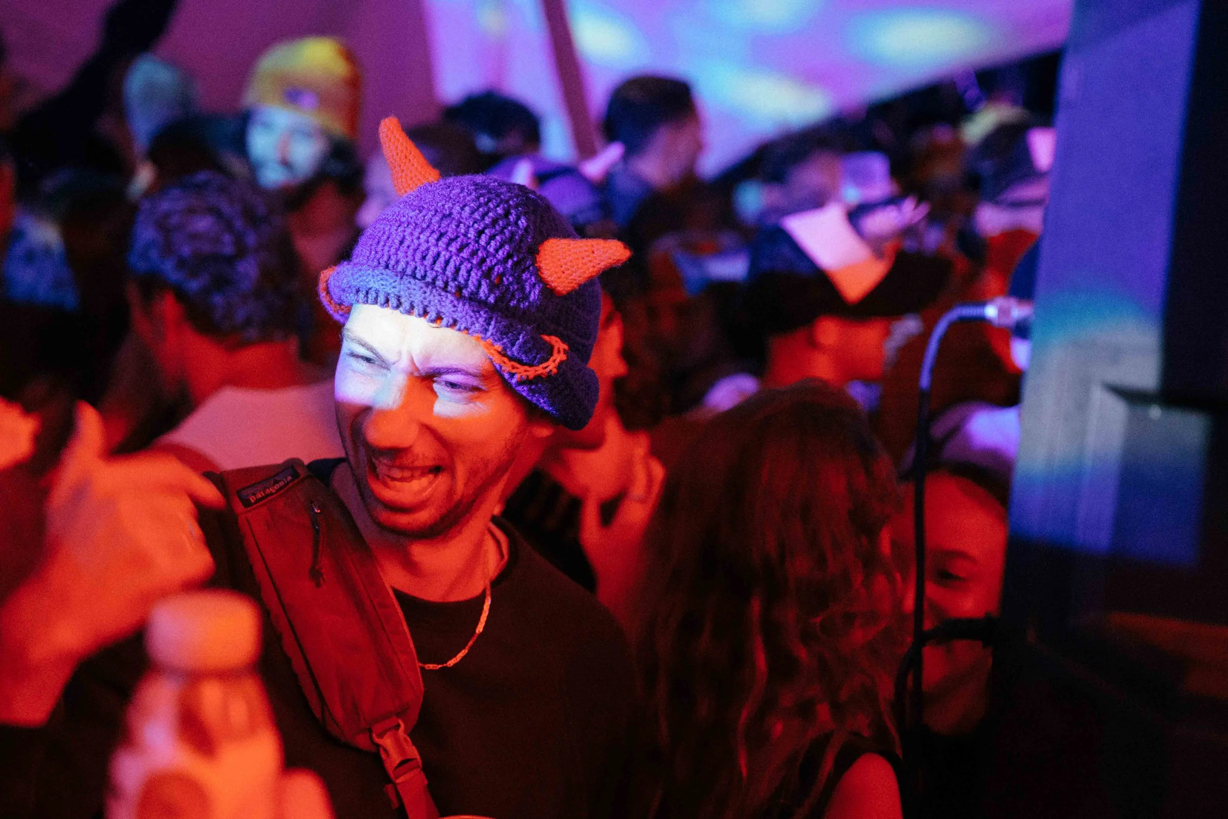 A man with a playful expression wearing a purple knit hat with orange horns and a red backpack in a crowded indoor event with colorful lighting.