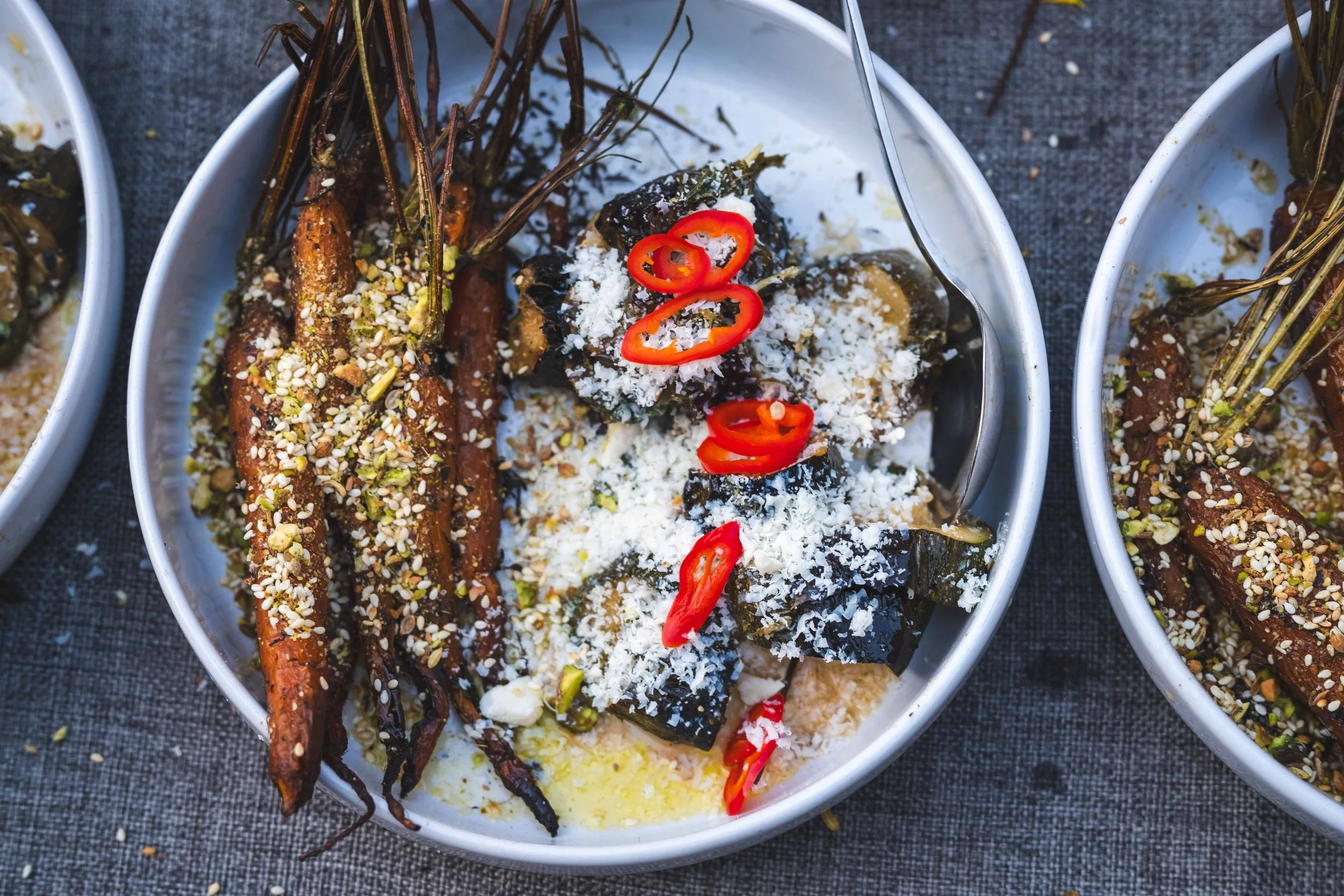 Bowl of roasted vegetables topped with red chili slices, grated cheese, and seeds on a dark cloth surface.