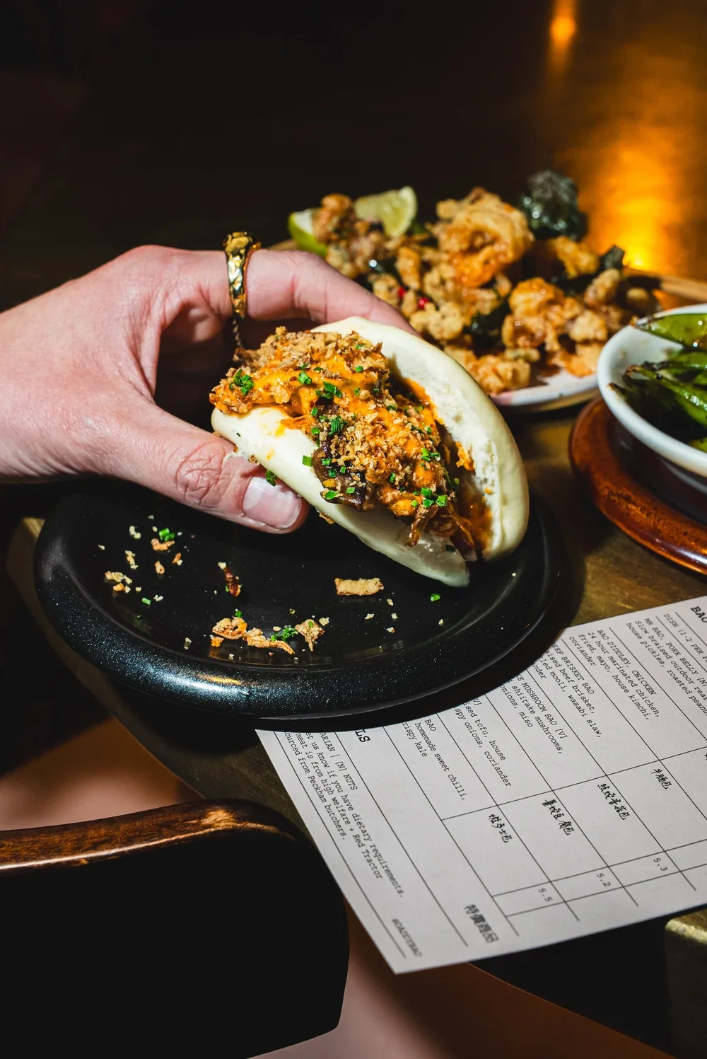 Hand holding a steamed bun filled with meat and garnished with chopped chives, with plates of fried food and a bowl of green chili peppers on a dark table.