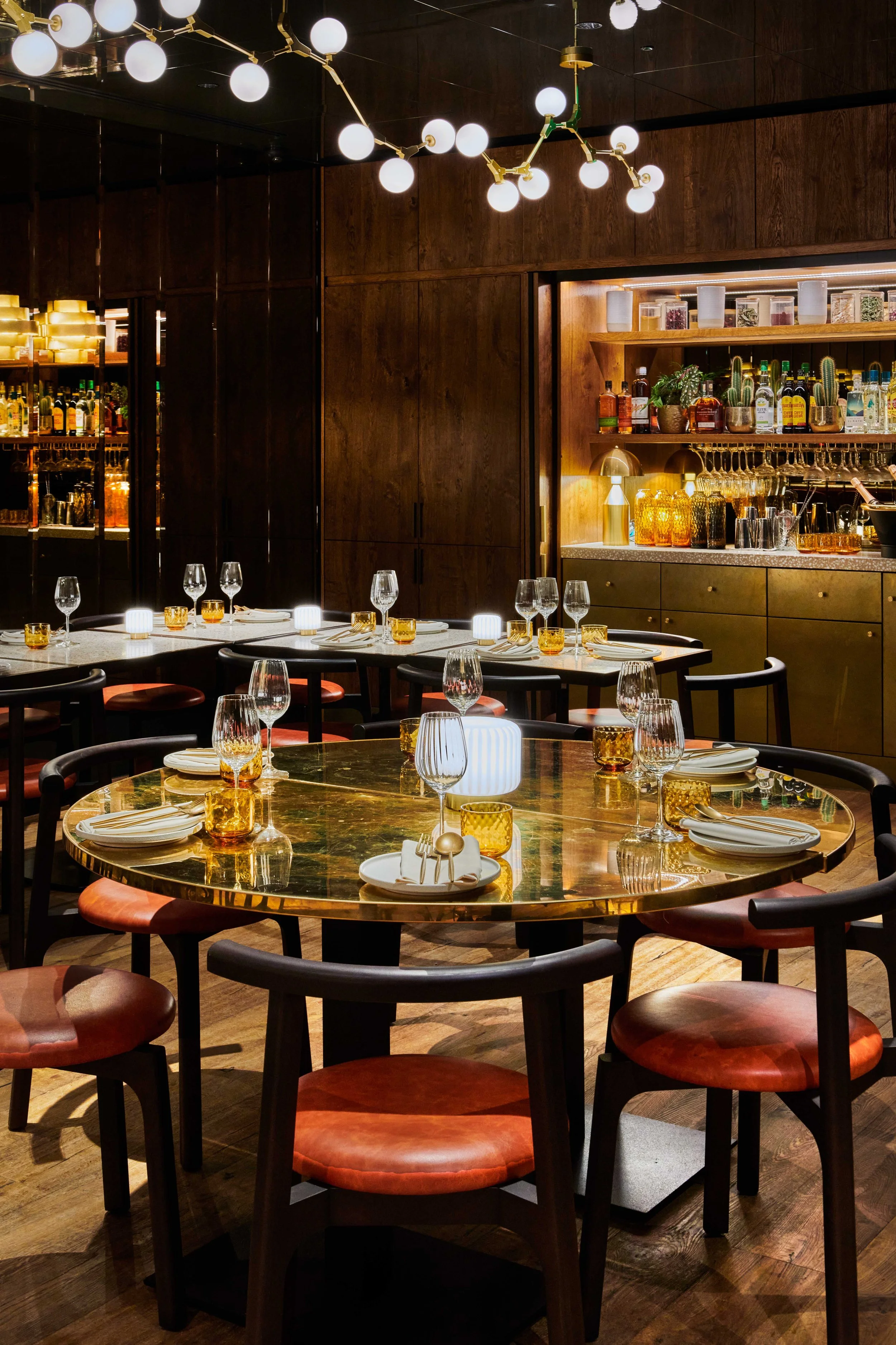 A dimly lit restaurant with round tables, set with glasses, plates, and cutlery, featuring a modern chandelier and a well-stocked bar in the background.