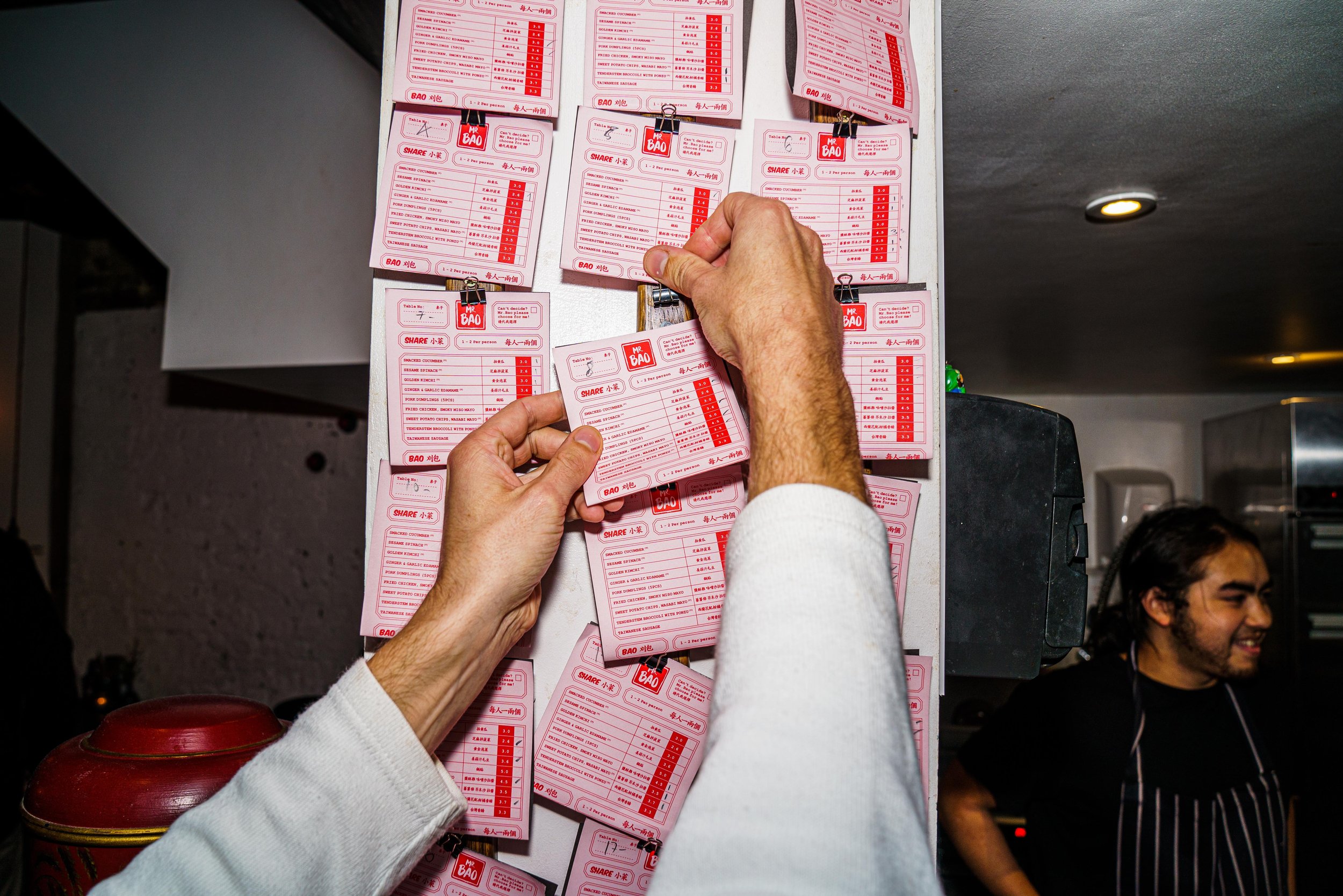 A person is pinning pink order slips on a wall, with a man in an apron smiling in the background inside a restaurant.