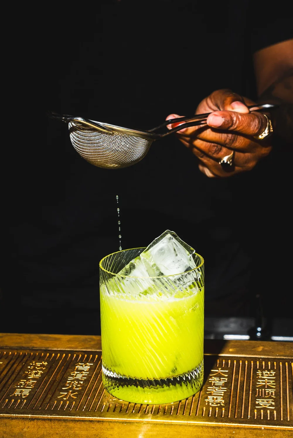 A person pours a liquid through a metal strainer into a glass with ice and a bright yellow-green beverage. The person wears rings and has dark skin. The background is black.