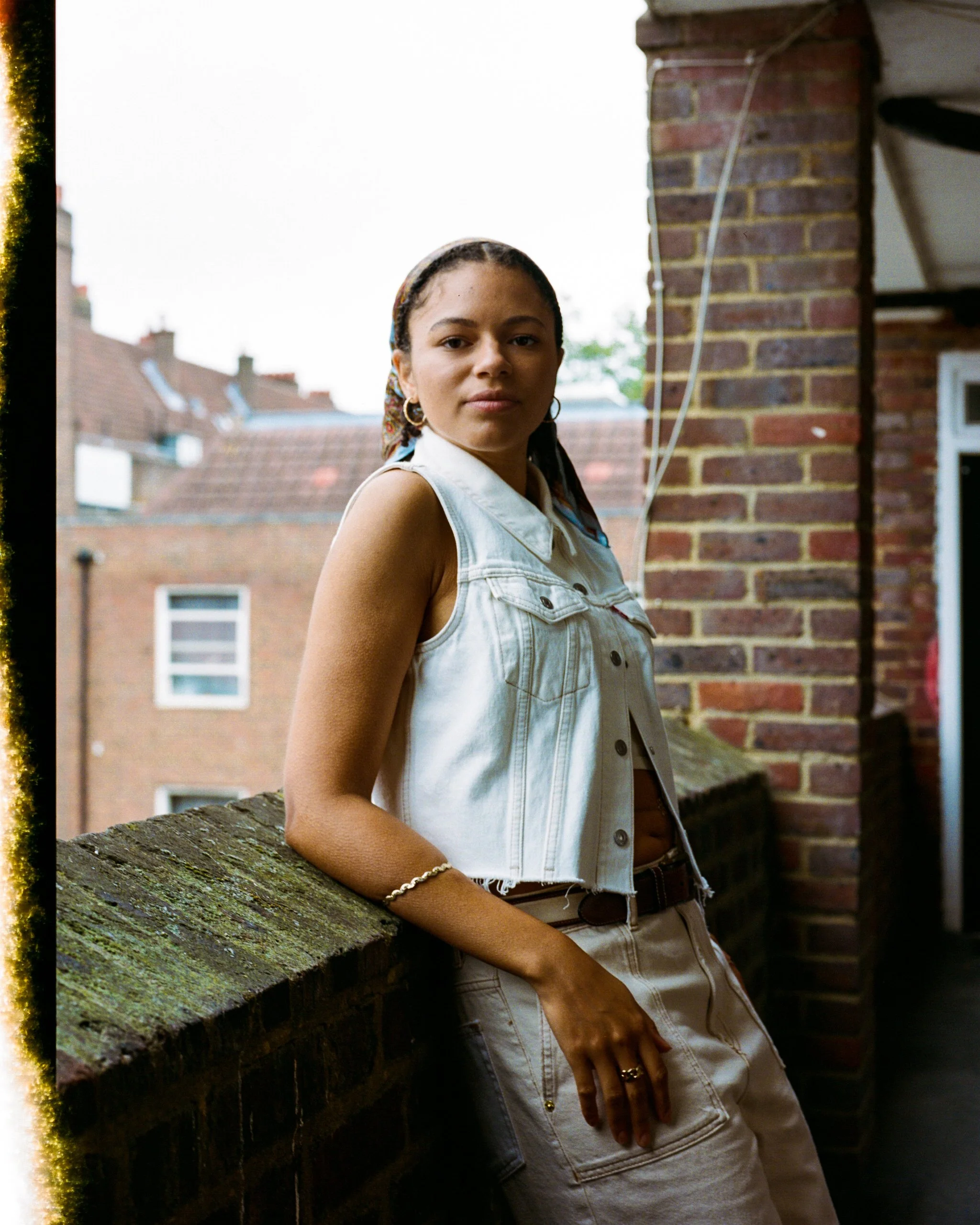 Young woman leaning against a brick balcony railing outdoors, wearing a sleeveless denim vest and beige cargo pants, with a background of houses and overcast sky.