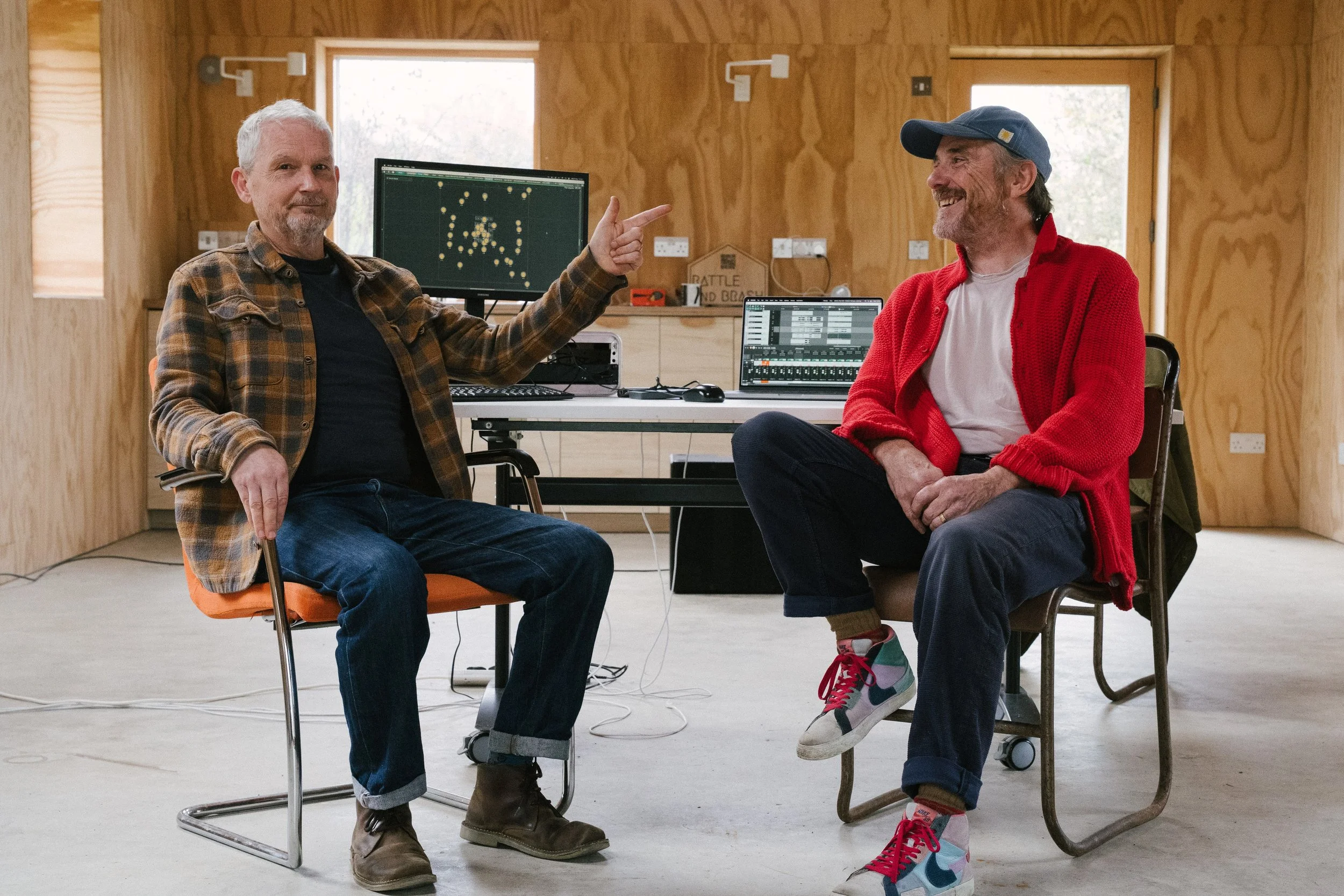 Two men with grey hair sitting in chairs in a wooden room, smiling, with one pointing at the other. There are computer screens behind them showing digital audio workstations and a tennis match.
