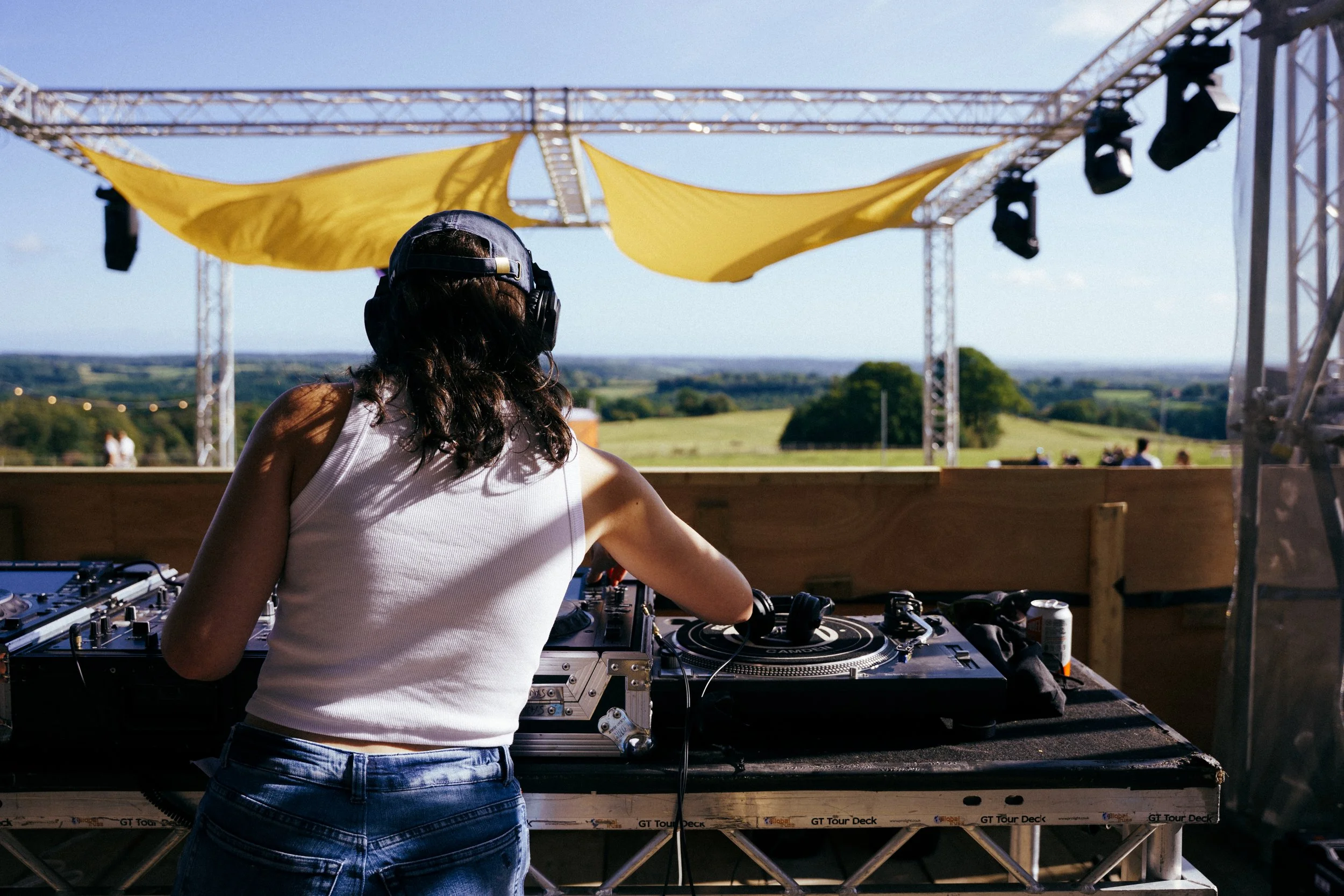 A female DJ performing outdoors on a sunny day, wearing a white tank top, headphones, and jeans, with DJ equipment on a table in front of her, and a landscape with green fields and a blue sky in the background.