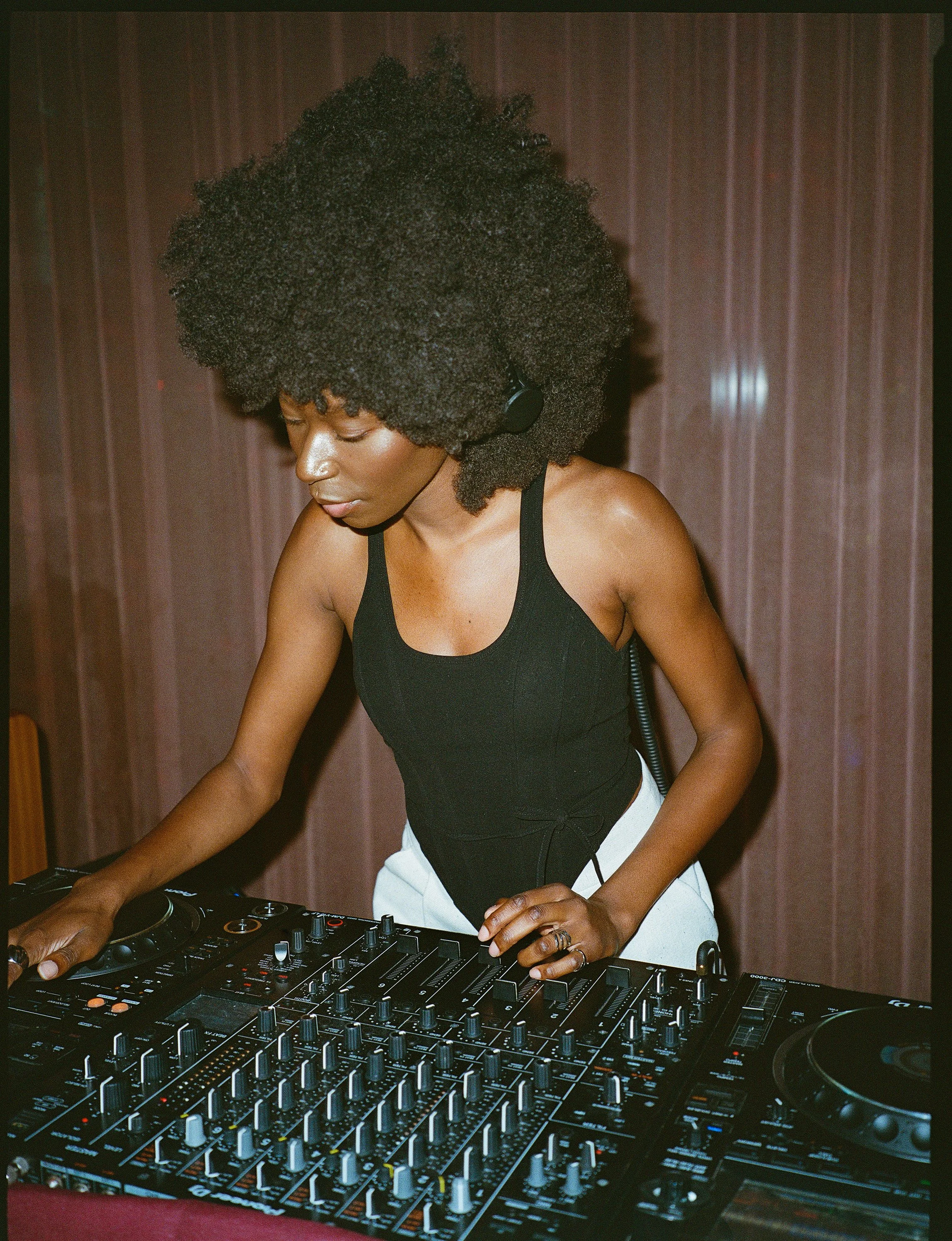 A woman with curly afro hair wearing headphones operating a DJ mixer in a dimly lit room.