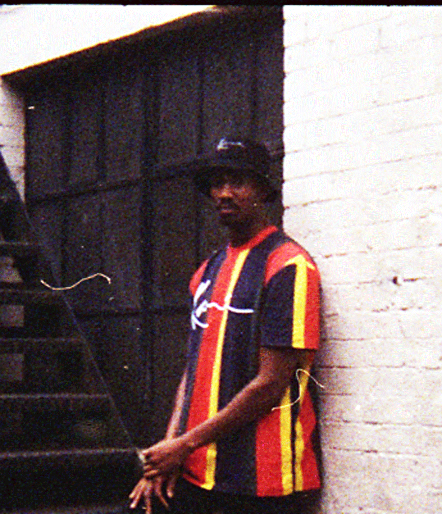 A person standing near a staircase against a white brick wall, wearing a colorful striped shirt, a black hat, and a silver chain.