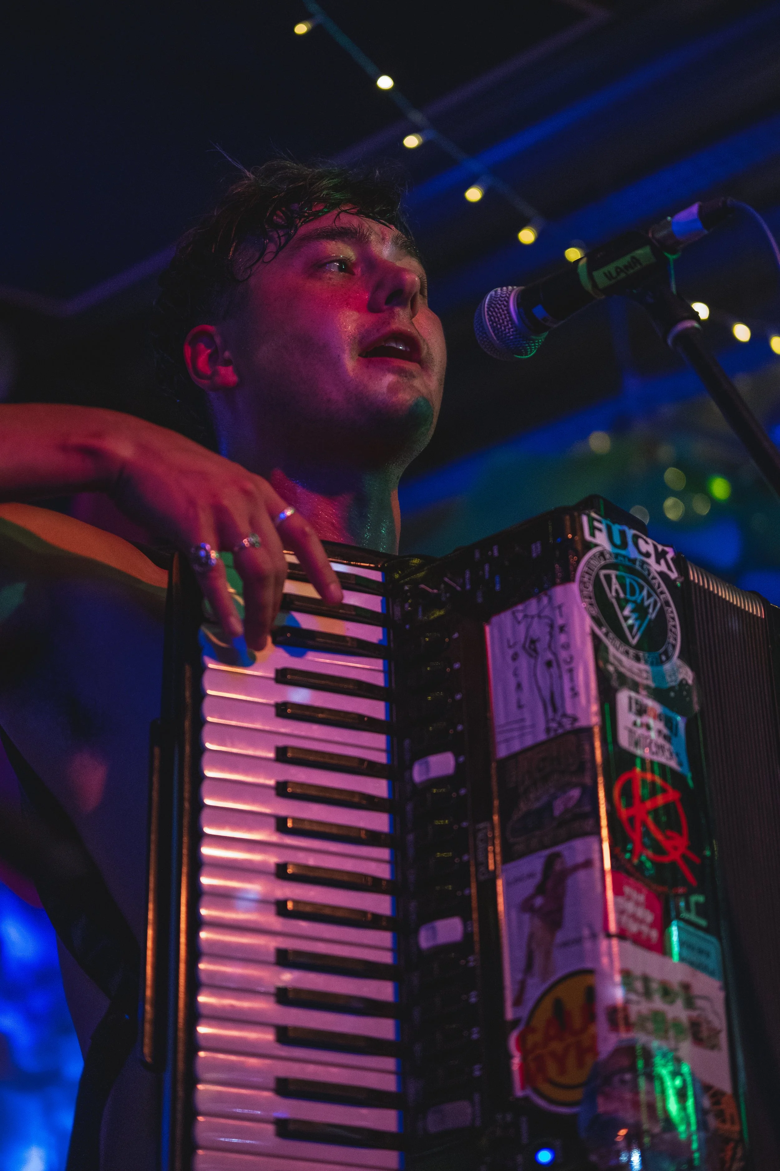 A young man performing on stage with colorful lighting, playing an accordion covered with stickers, and singing into a microphone.