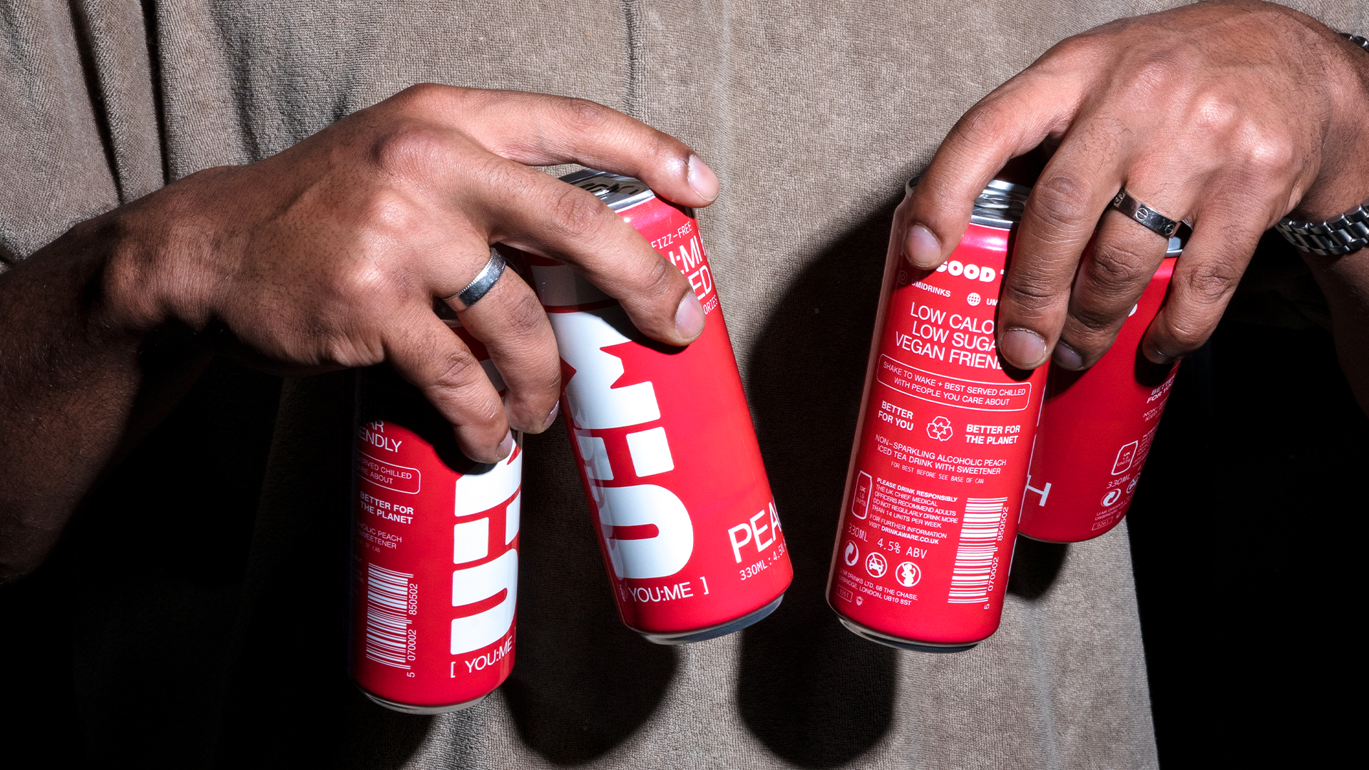 Two hands holding cans of a low-calorie, vegan-friendly beverage with red packaging and white text.