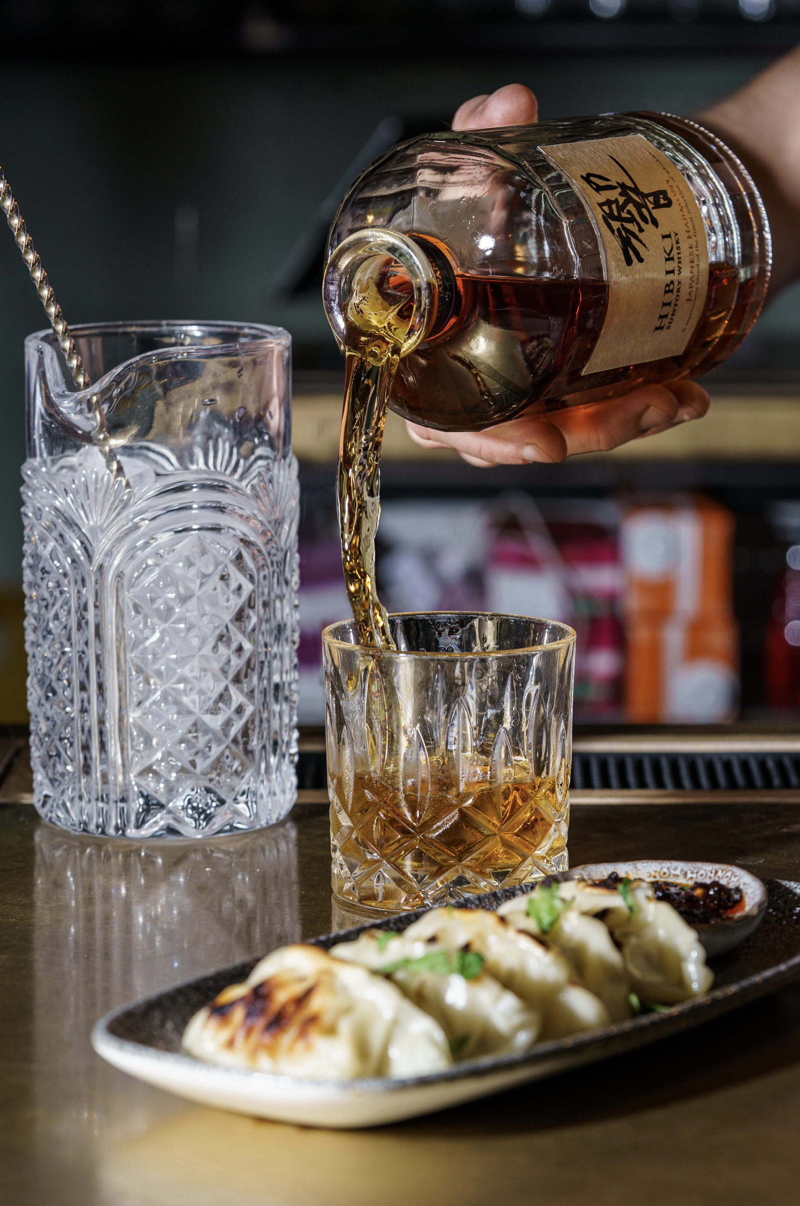 A hand pouring whiskey from a bottle into a glass on a bar counter, with a plate of dumplings and a small dish of dipping sauce in the foreground.
