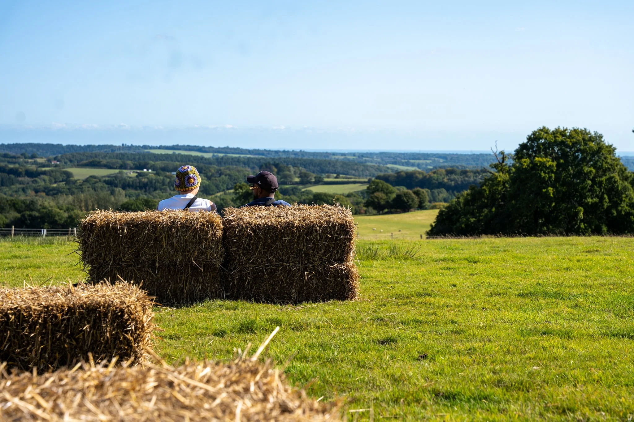 Two people sitting on hay bales in a green field, overlooking rolling countryside and trees under a clear blue sky.