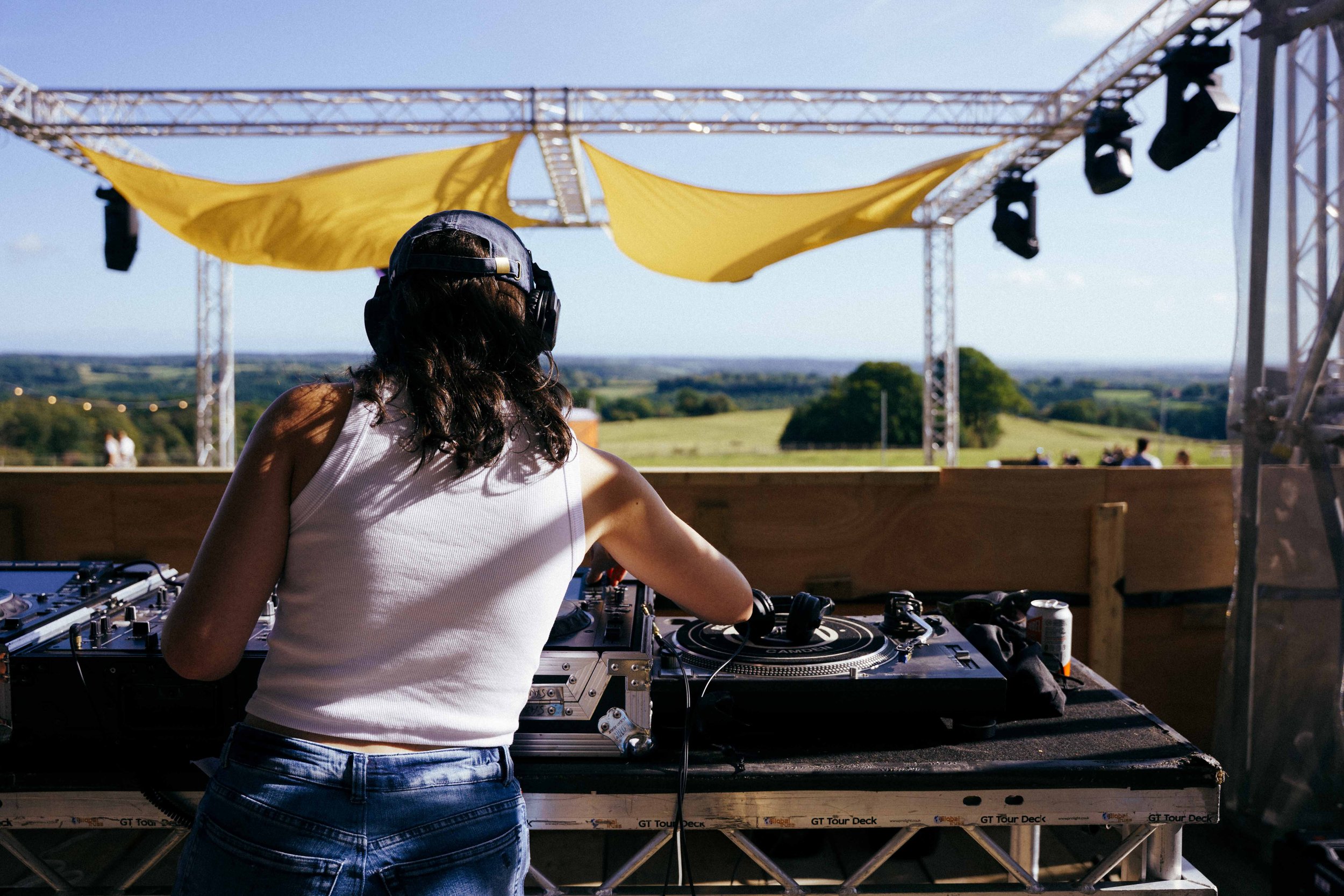 A female DJ wearing a white tank top and denim shorts wearing headphones, DJing outdoors on a stage with a landscape of green fields and trees in the background. The stage has yellow fabric sunshade and black stage lights.