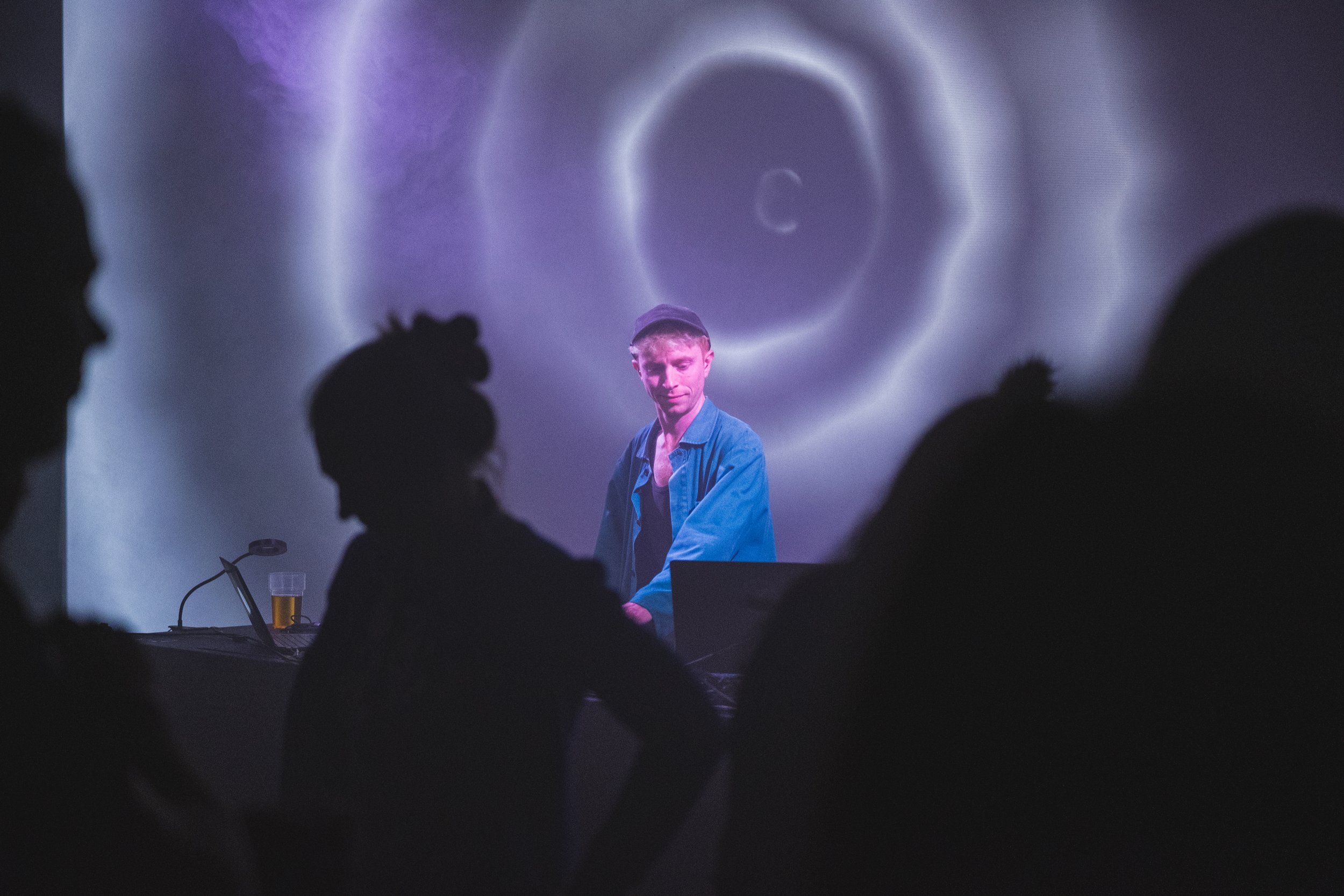 A DJ performs on stage with a swirling light pattern in the background, and silhouettes of audience members in the foreground.
