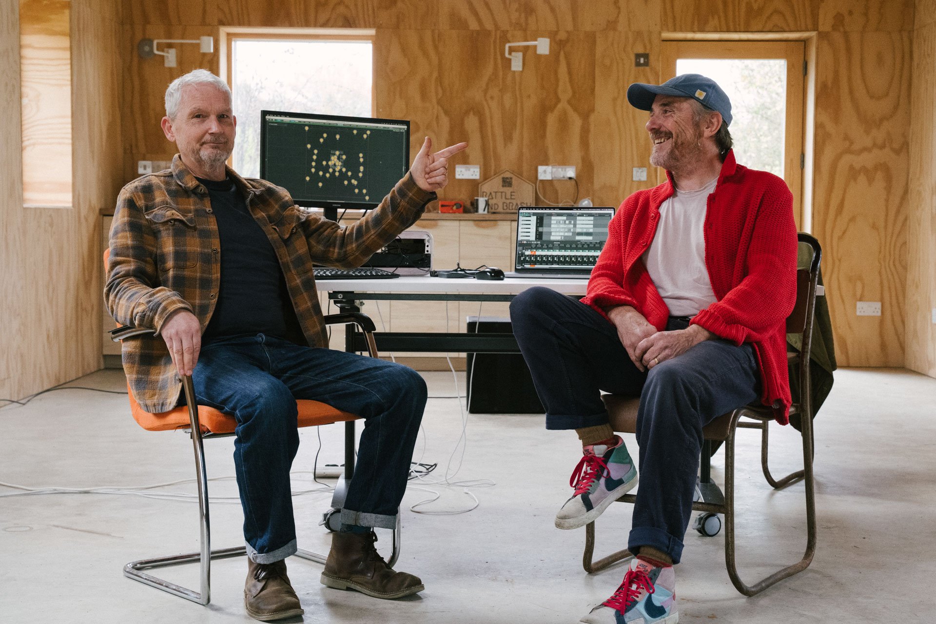 Two men sitting and talking in a wooden room with computers and screens in the background.