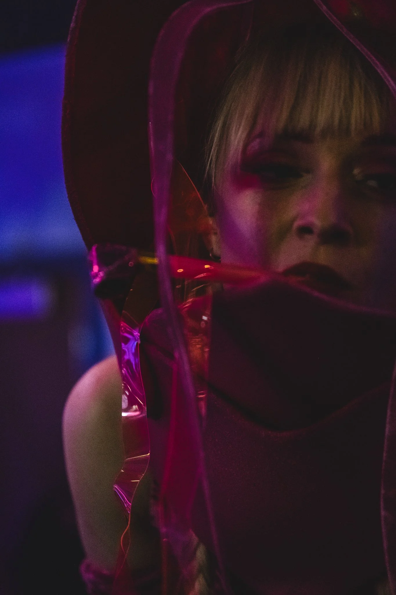 A person with blonde hair and makeup looking into a reflective, pink-tinted surface in a dark environment with colorful lighting.