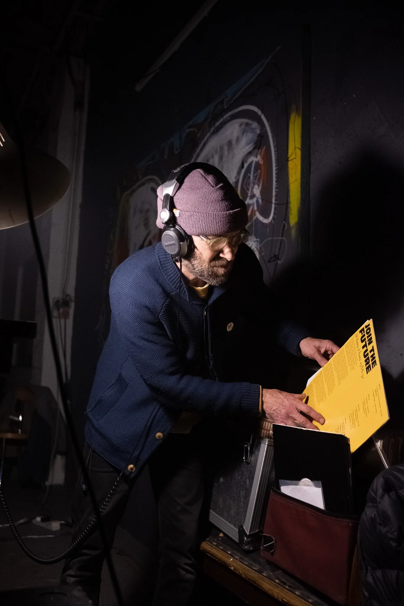 A man wearing glasses, a beanie, and headphones is reading a yellow poster or sheet in a dark room.