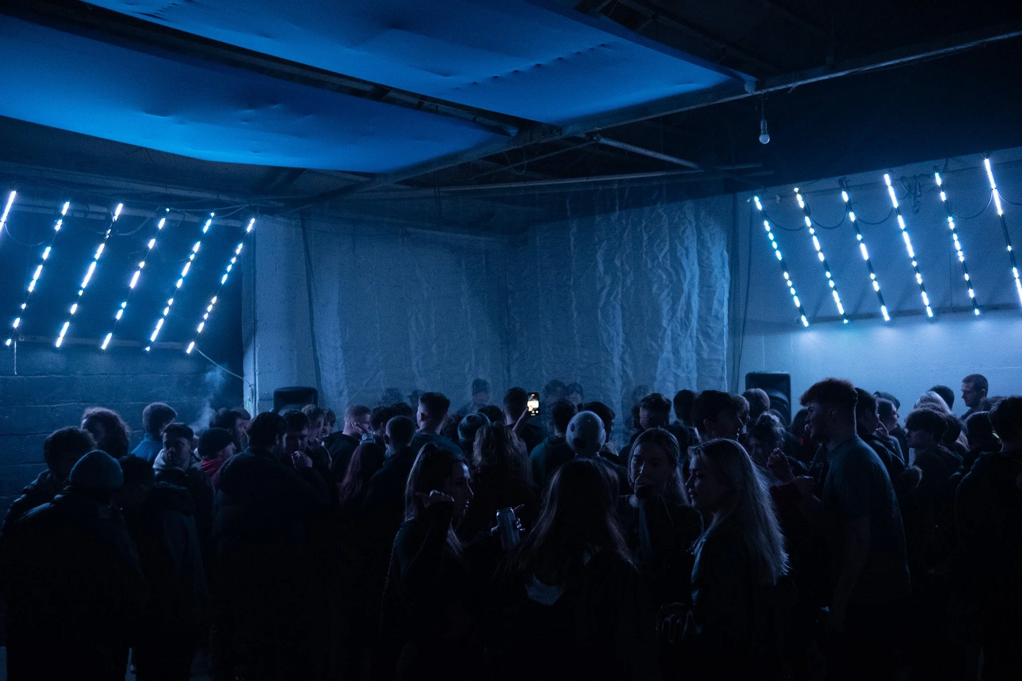 A crowded indoor event with people gathered together in a dimly lit space, illuminated by blue LED strip lights on the walls.