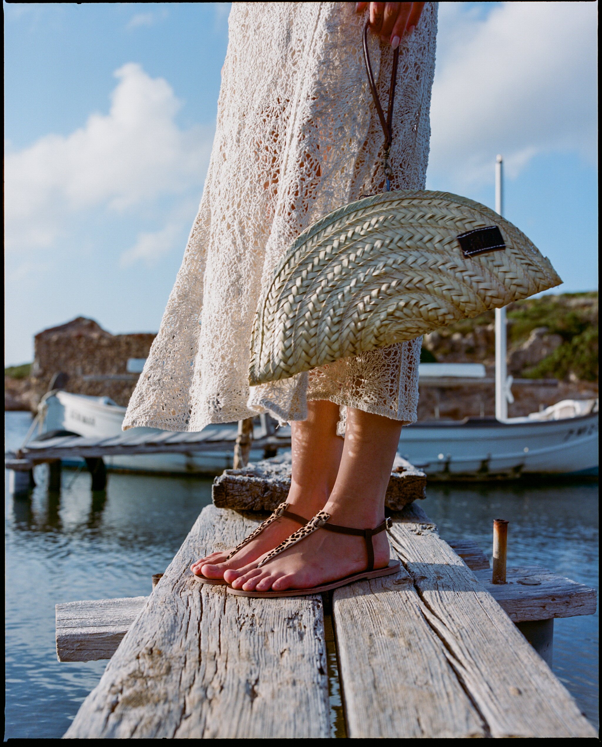 Person standing on a wooden dock wearing barefoot sandals, holding a woven straw clutch bag, near boats and water under a partly cloudy sky.