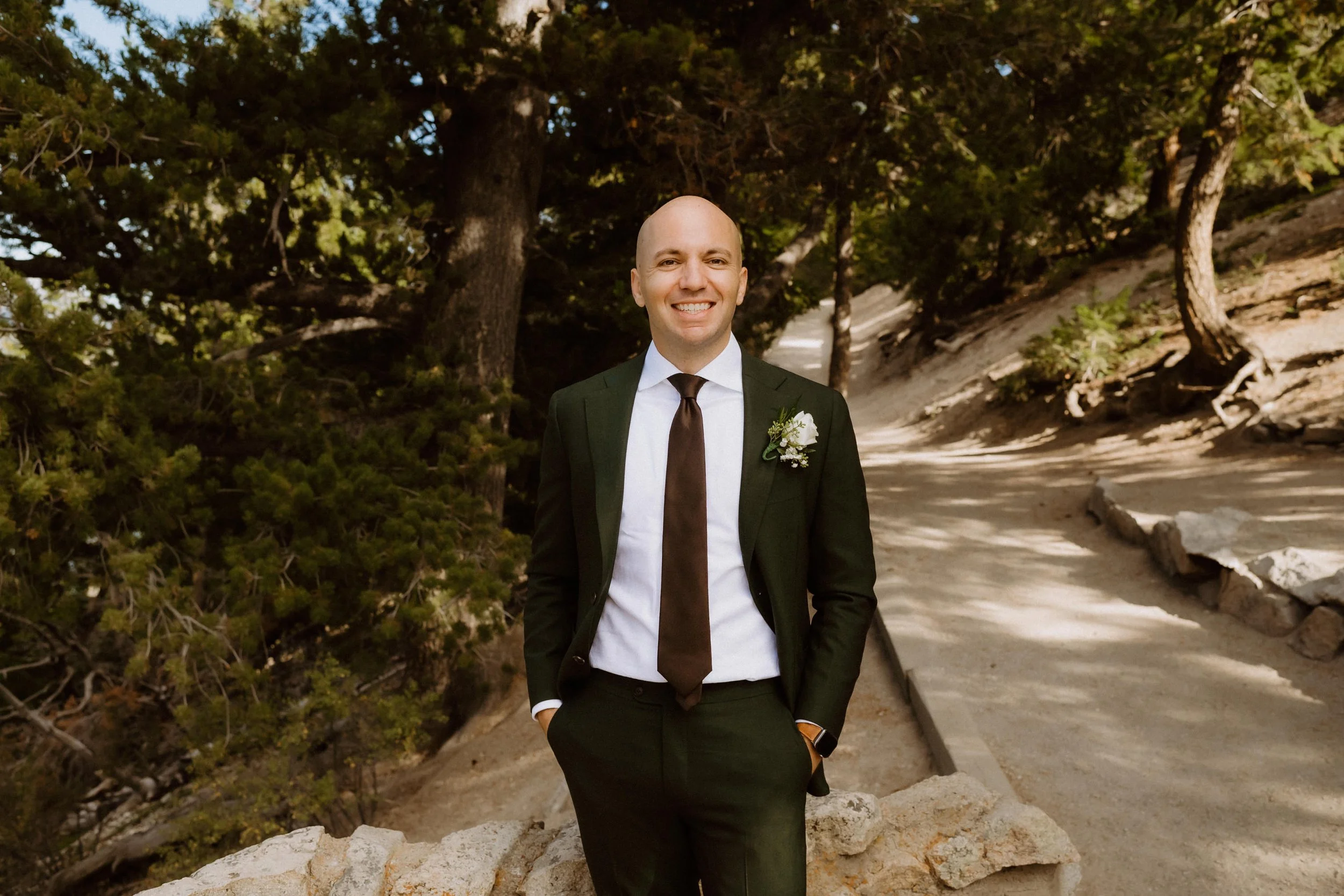 A man in a black suit with a white shirt and brown tie standing outdoors on a sandy path with trees in the background, smiling and hands in his pockets, wearing a white boutonniere on his lapel.