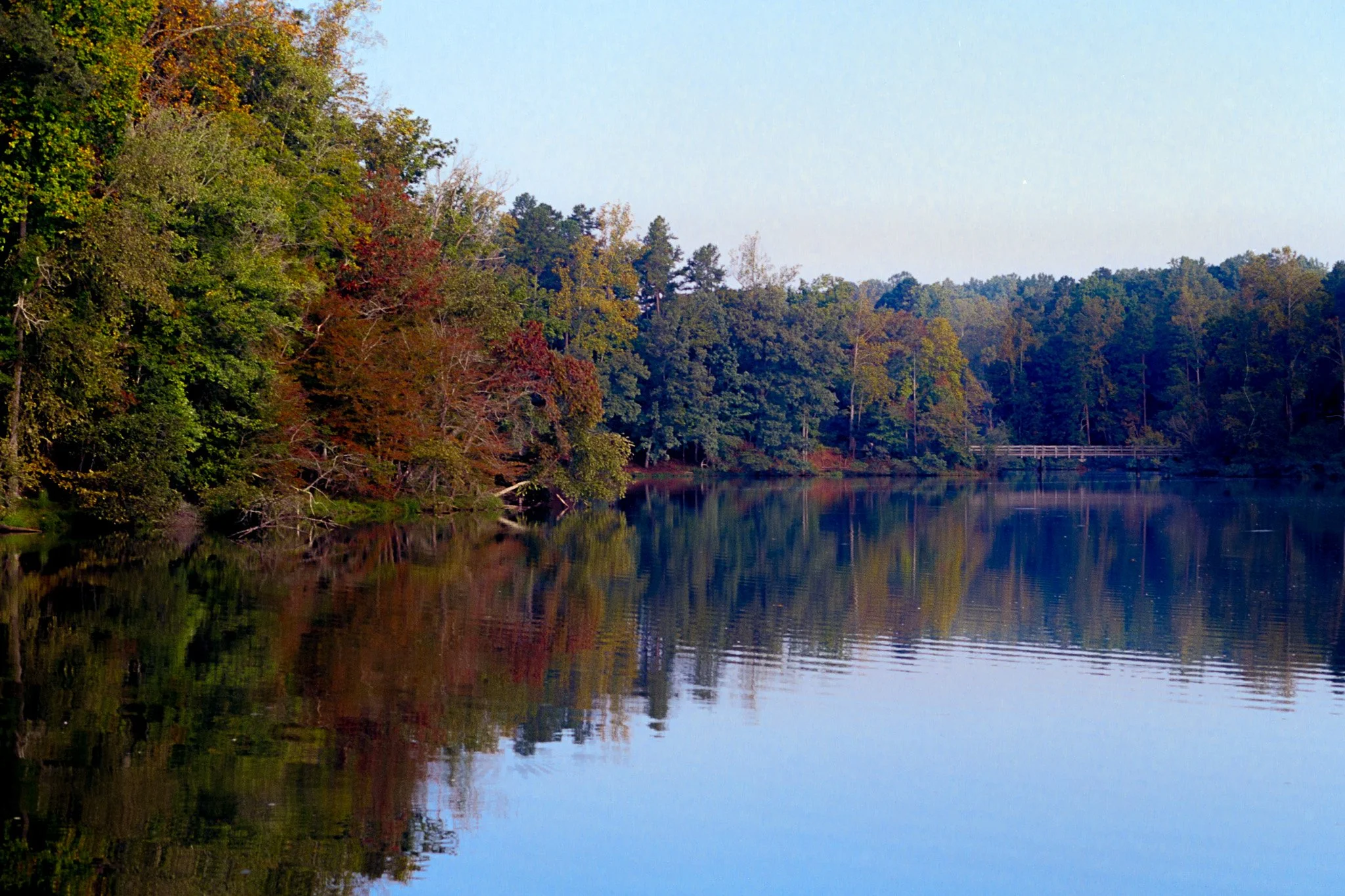 A calm river or lake reflecting trees with fall foliage along the banks, with a small bridge visible in the distance.