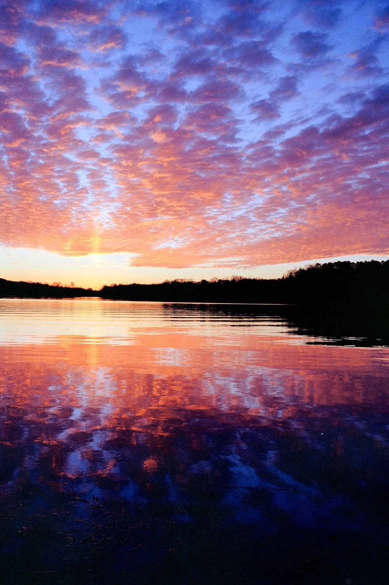 Sunset over a calm lake with pink and purple clouds in the sky, reflected on the water's surface.