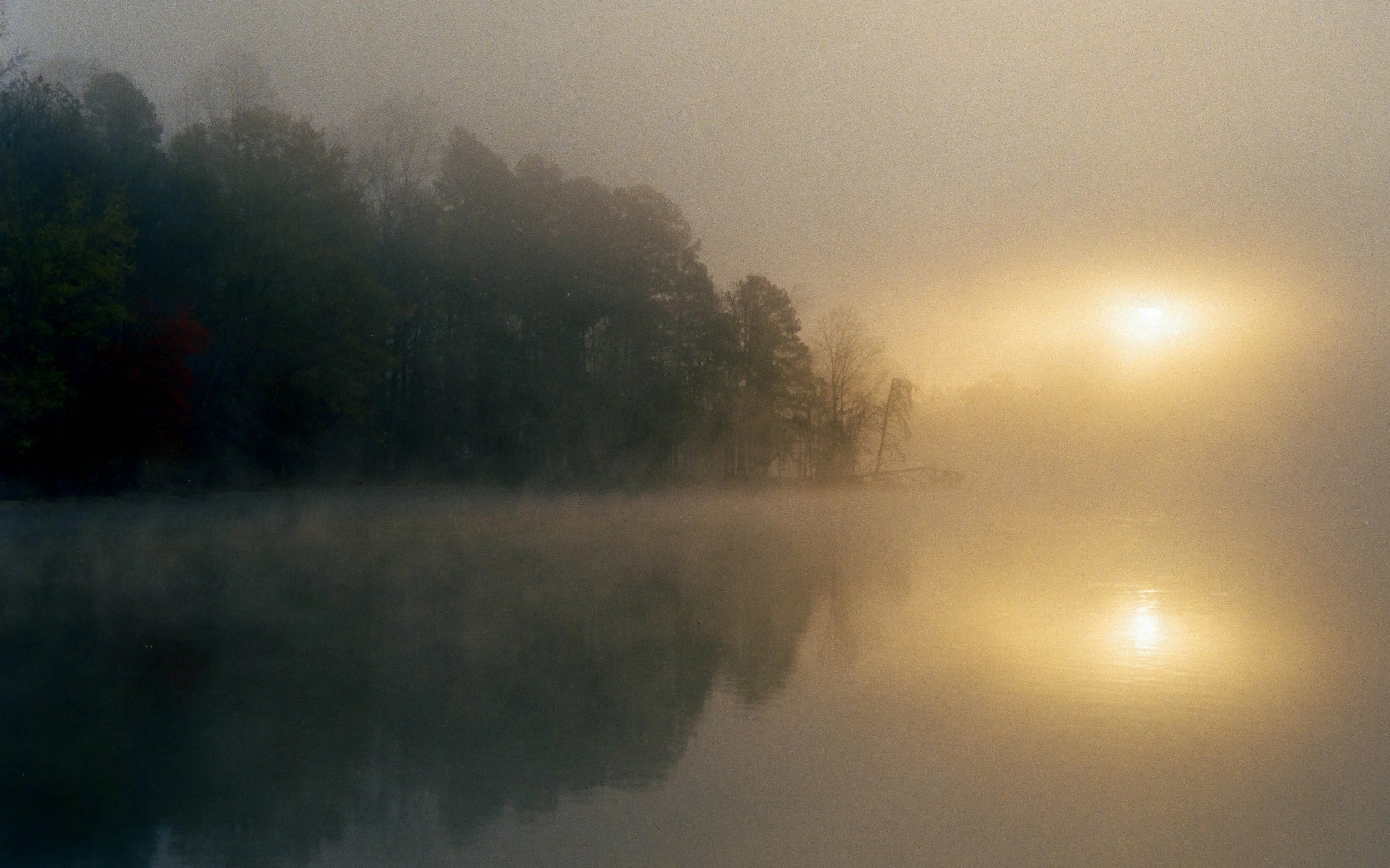 Early morning fog over a calm body of water with trees in the background and the sun partially obscured by the mist.