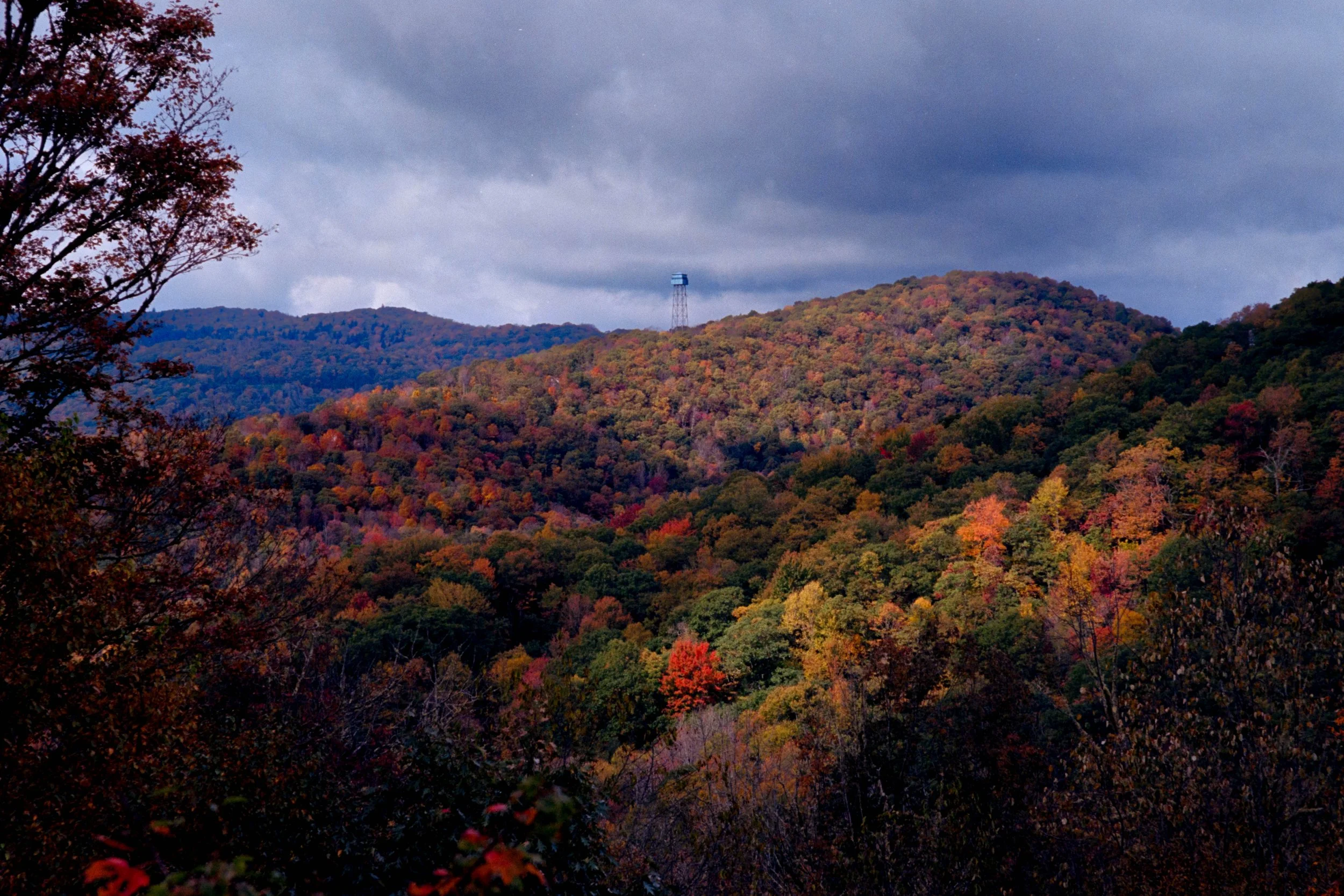 A panoramic view of a mountain covered in colorful fall foliage under a cloudy sky, with a tower visible on the ridge.