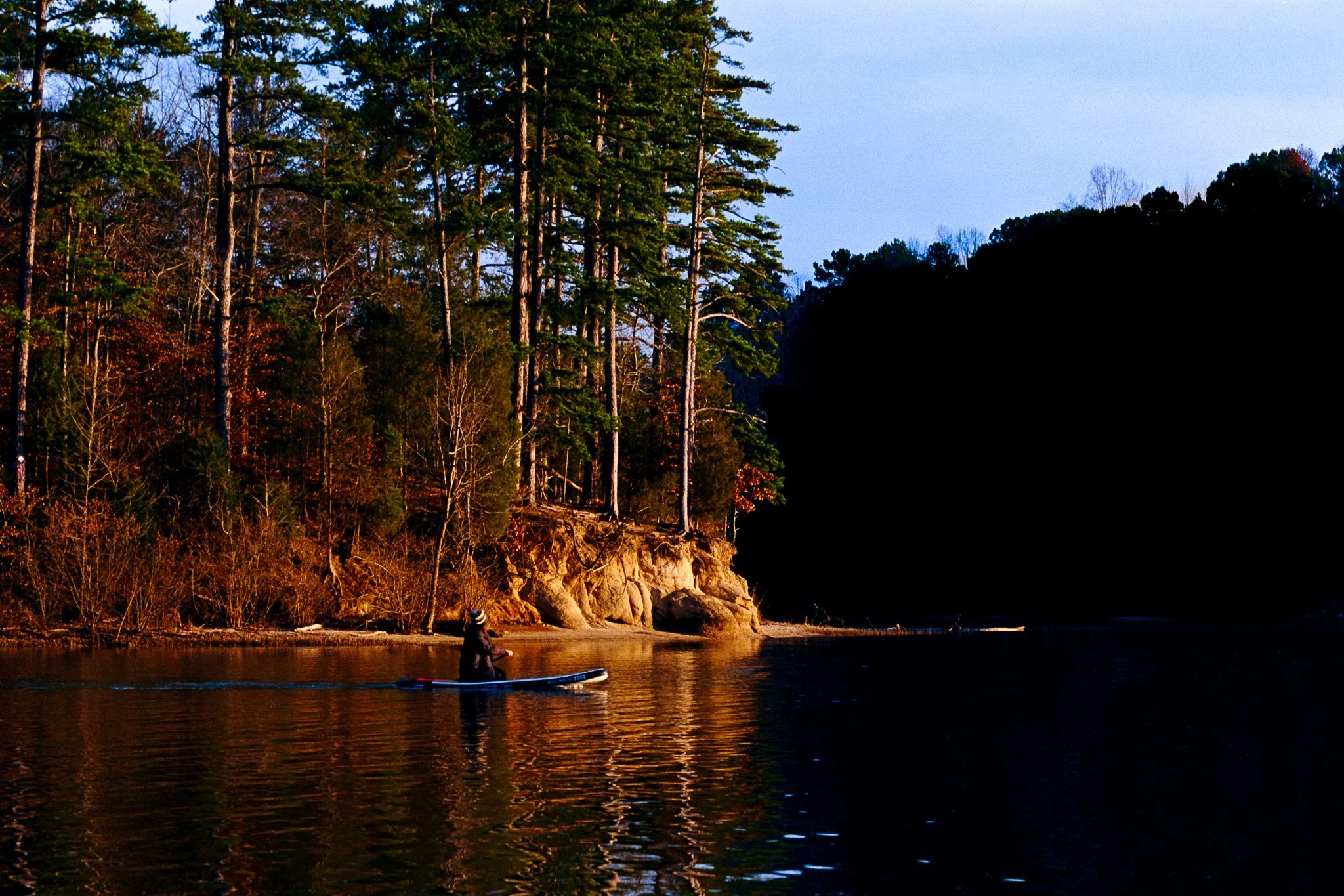 A person standing on a paddleboard on a calm river near a shoreline with tall trees and rocky cliffs, during late afternoon or early evening.