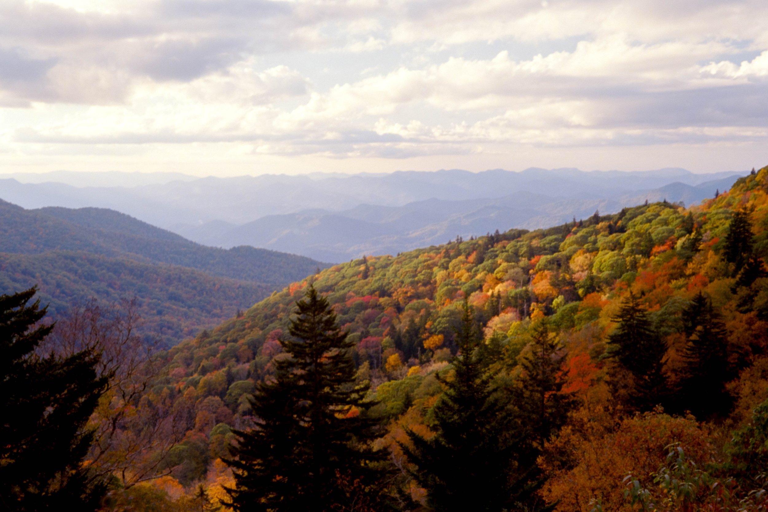 A scenic view of a mountain range with forested hills and trees displaying fall colors, under a partly cloudy sky.