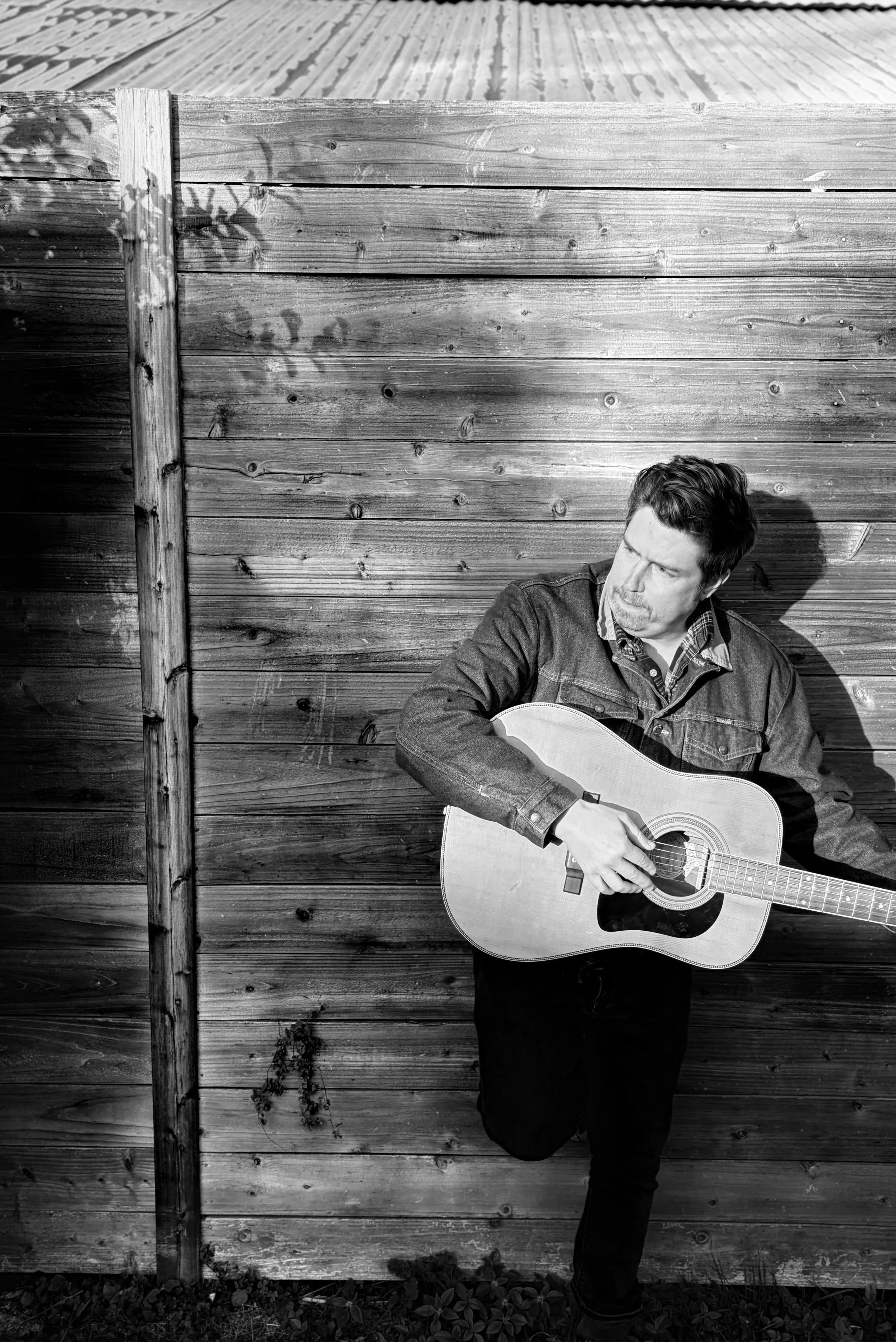 A man in a denim jacket playing an acoustic guitar outdoors against a wooden fence, with shadows and sunlight highlighting the scene.