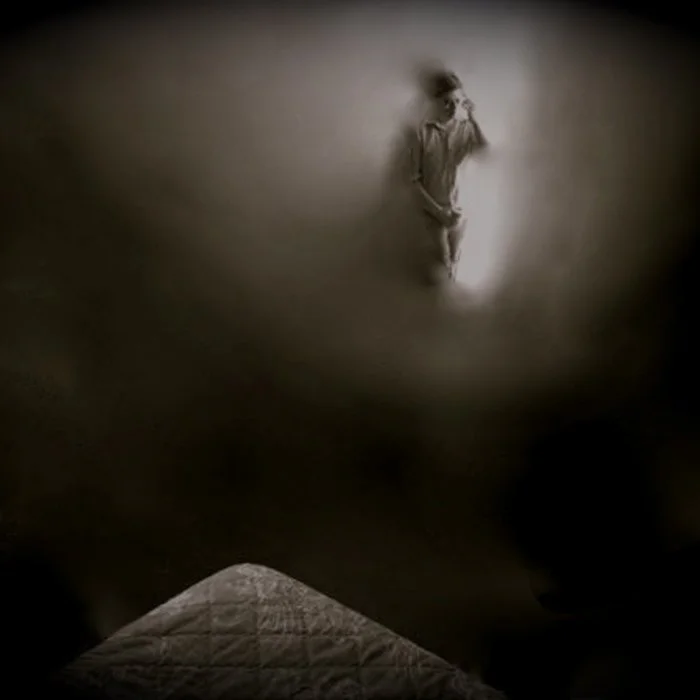 Man dressed in light-colored casual clothing, walking in a dimly lit corridor with a single light source, casting shadows on the wall.