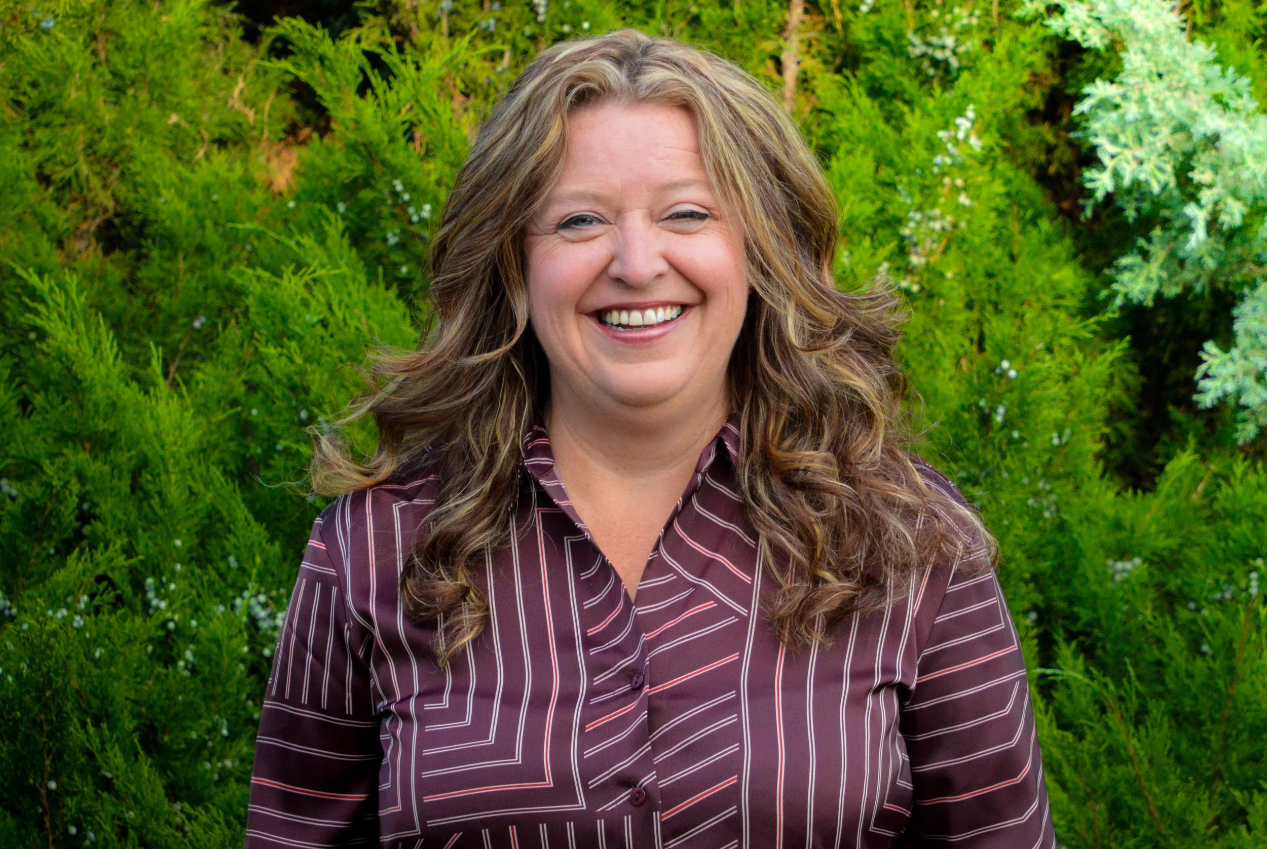A cheerful woman with wavy, shoulder-length blond hair wearing a maroon and white striped button-up shirt, standing outdoors in front of green shrubbery.