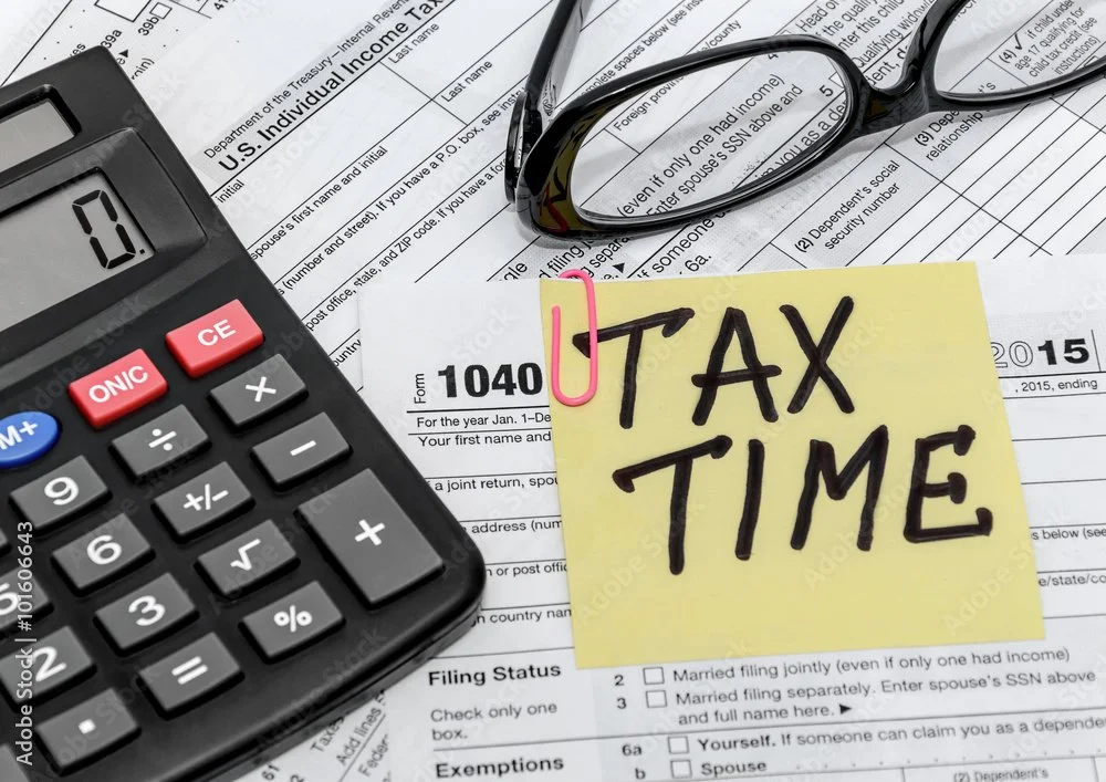 A calculator, a pair of reading glasses, a handwritten note saying 'TAX TIME', and several tax forms and documents on a desk.