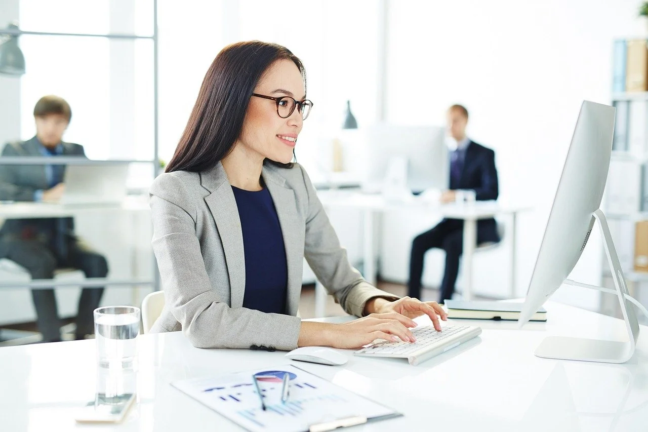 Woman in business attire working at a computer in an open office with colleagues in the background.