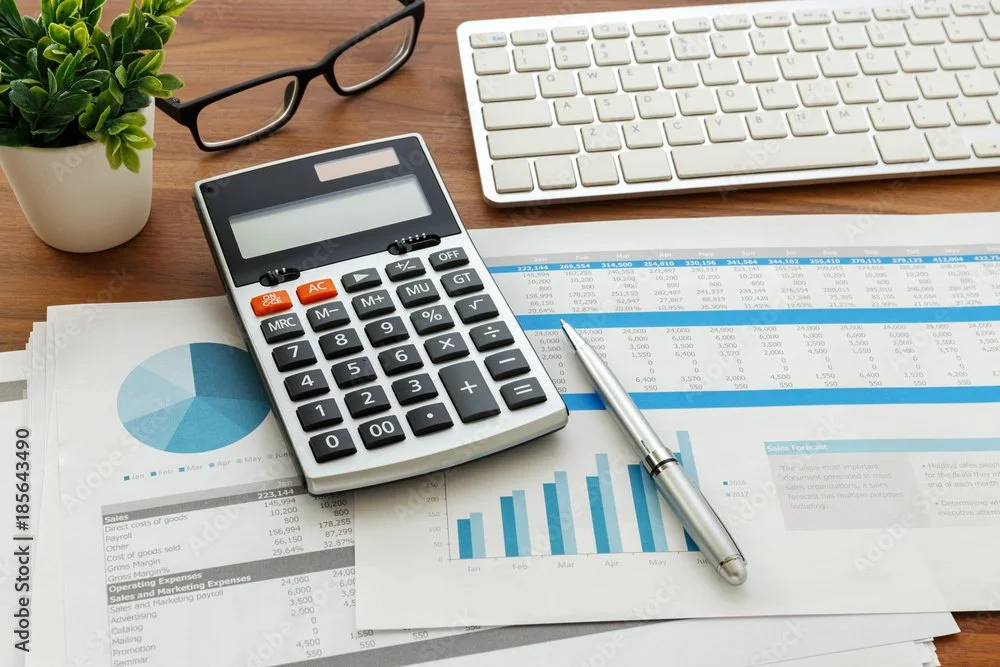 Desk with a calculator, a pen, a spreadsheet, and a graph, along with a keyboard, glasses, and a potted plant.