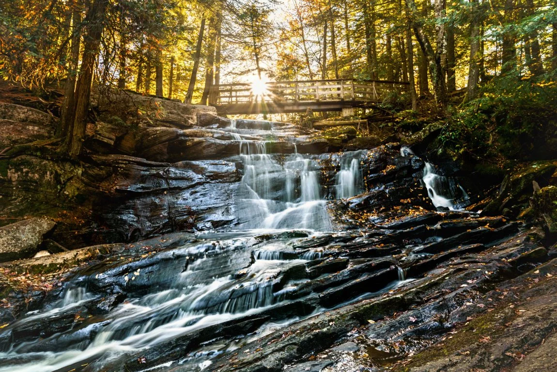 A cascading waterfall flowing over black rocks in a forest, with sunlight shining through the trees and a wooden bridge at the top.