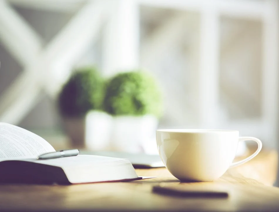 A white coffee cup, an open book, and a pen on a wooden table with blurred potted plants in the background.