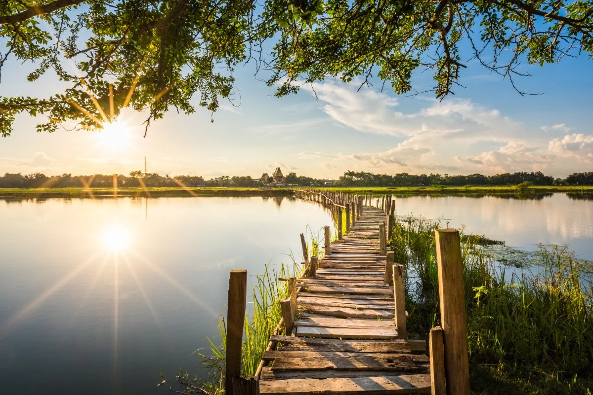 A wooden dock extends over a calm lake at sunset, with trees and a traditional building in the distance.