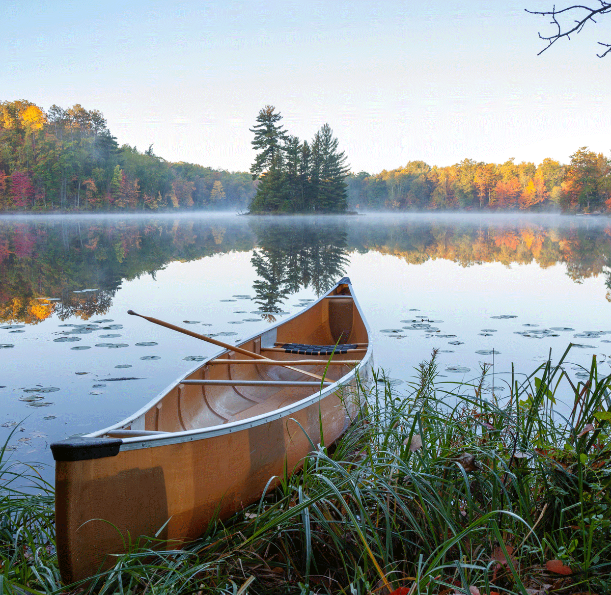 A canoe resting at the edge of a calm lake during autumn, with trees displaying fall colors, and a small island with tall trees in the background, reflecting on the water.