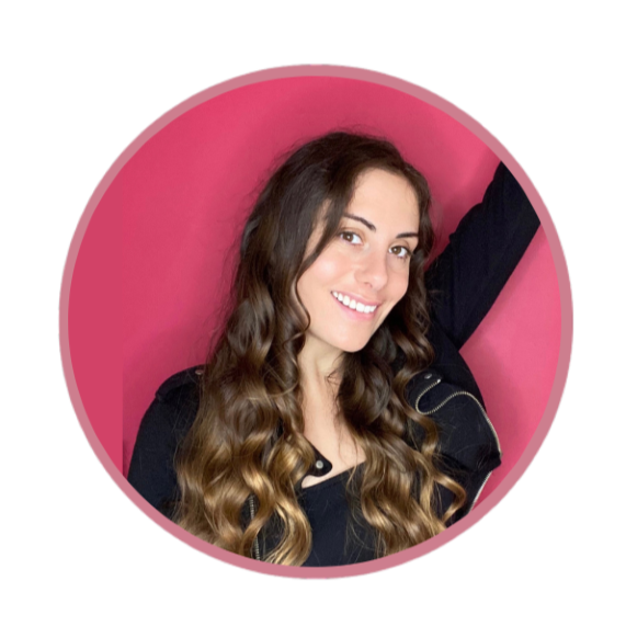 A smiling woman with long wavy hair taking a selfie against a pink background, wearing a black top.