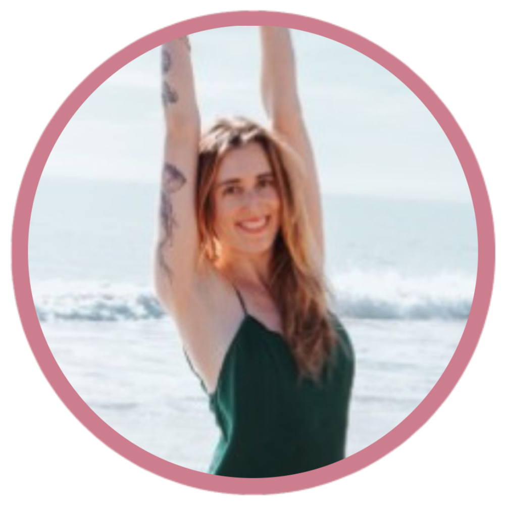 A woman with long red hair and tattoos on her arms standing on the beach with her arms raised above her head, smiling at the camera, with the ocean and sky in the background.