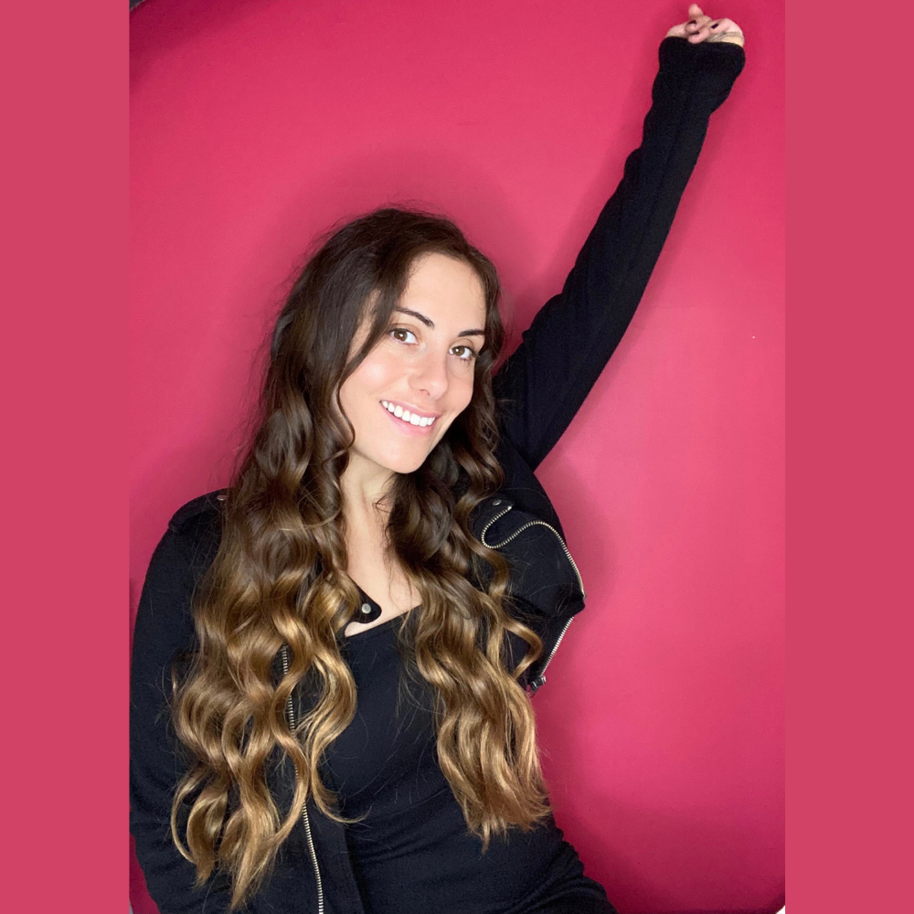 Person with long wavy hair smiling and raising one arm against a pink background