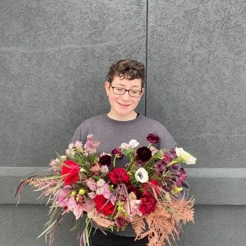 Person with short curly hair and glasses holding a large bouquet of colourful flowers