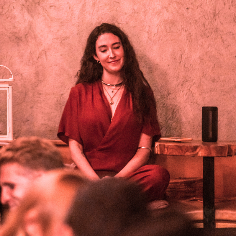 Curly-haired woman in a red dress sitting cross-legged indoors, smiling with eyes closed