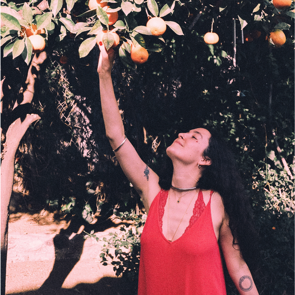 Isabella Frappier, a light skinned woman with long dark hair and a tattoo on her arm, reaches up to pick an orange from a tree. She is smiling.