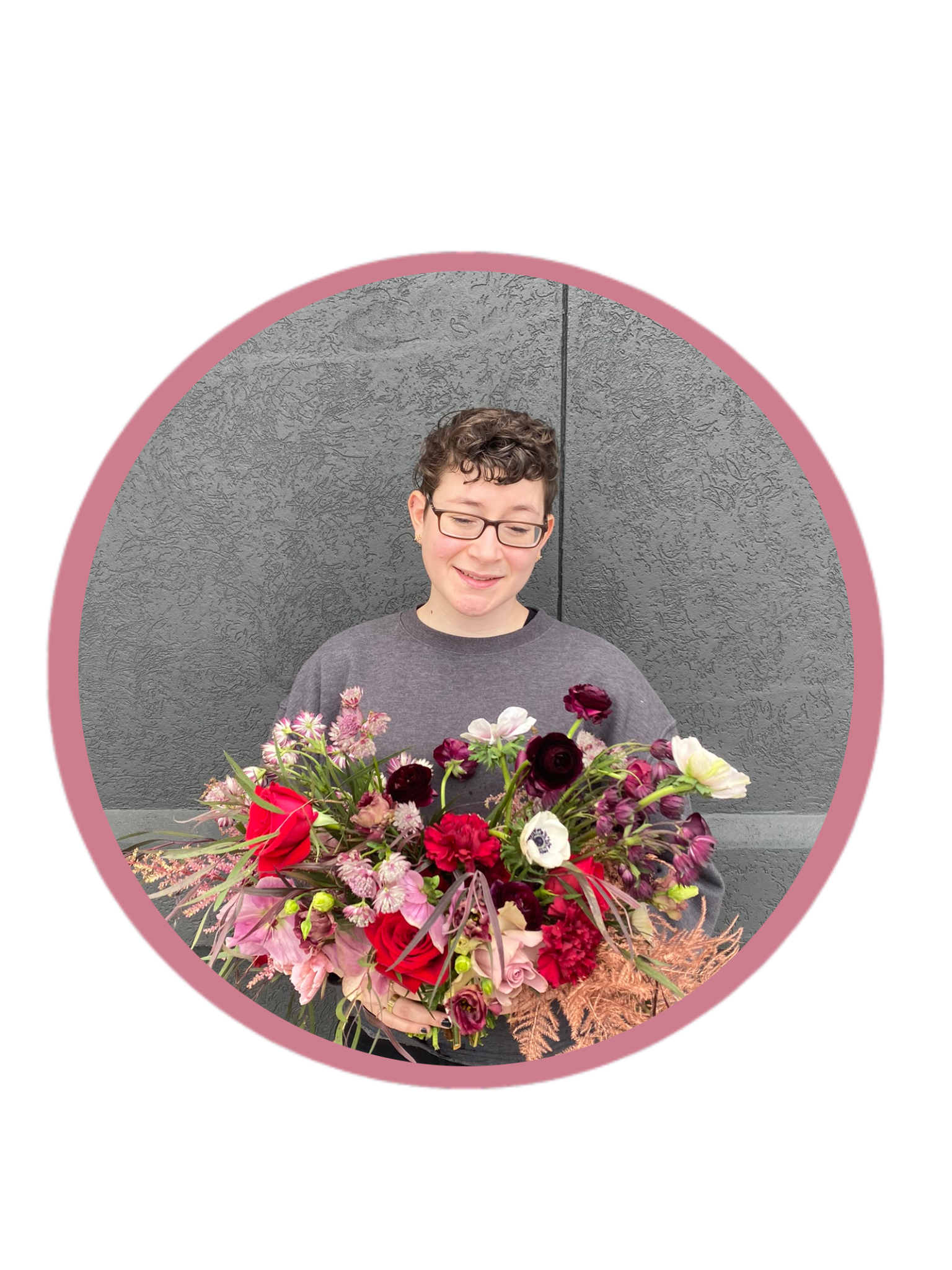 A person with short, curly hair and glasses holding a large bouquet of colorful flowers, standing against a gray textured wall.