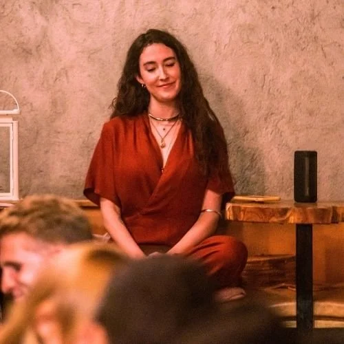 Curly-haired woman in a red dress sitting cross-legged indoors, smiling with eyes closed representing online personal development mentorship and coaching